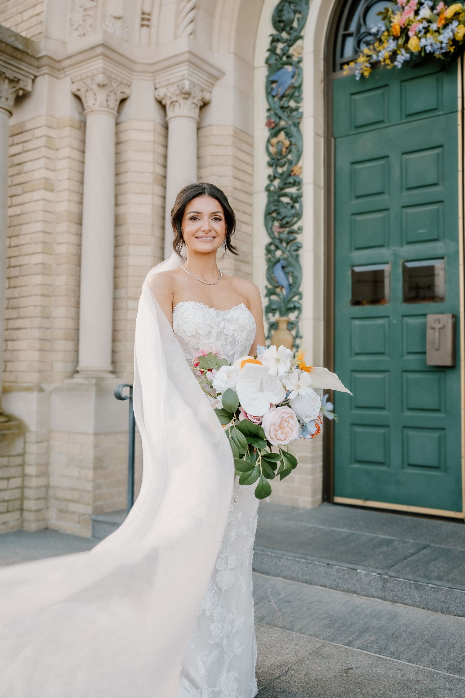 Smiling bride in strapless lace gown holding bouquet on church steps by teal door.