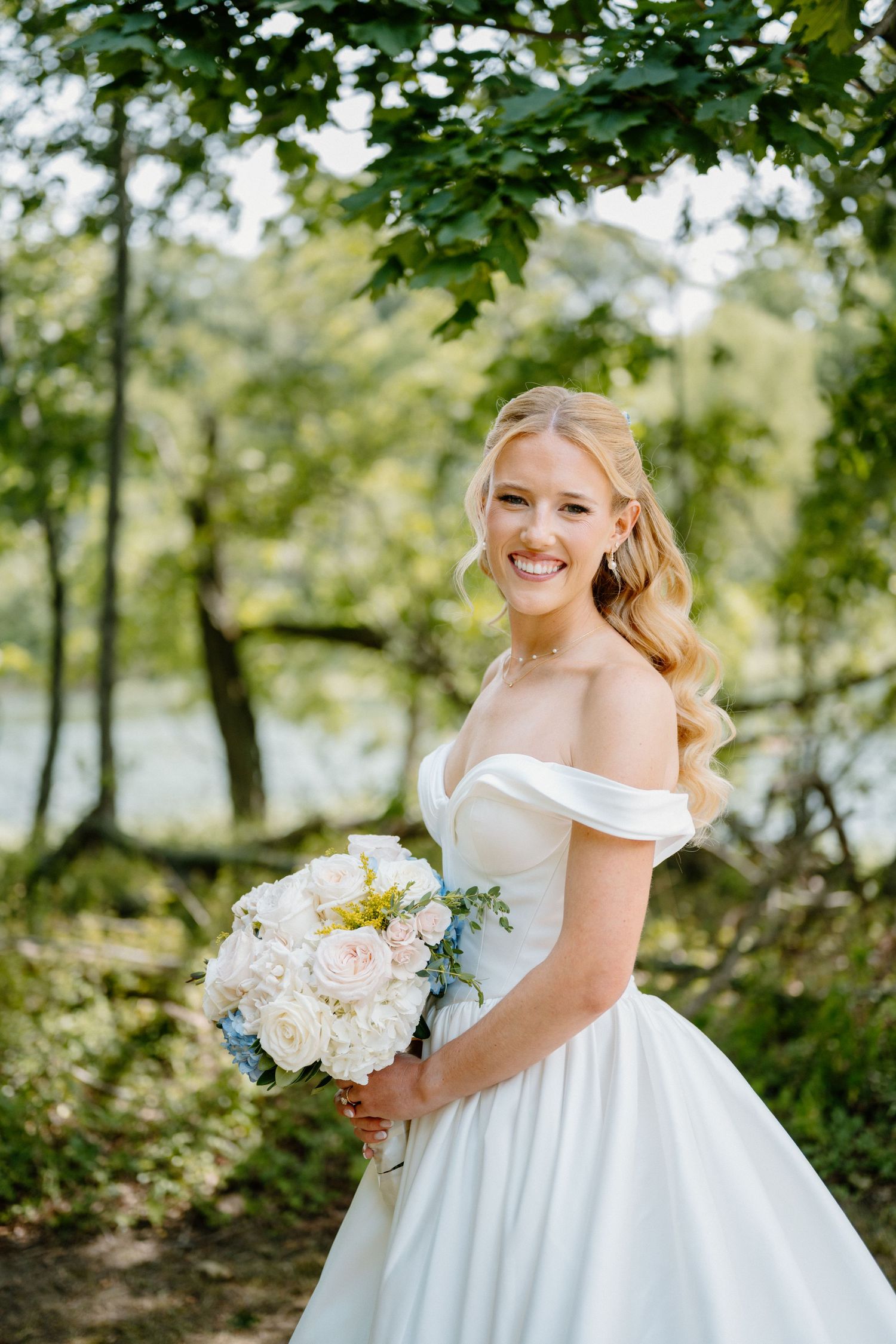 Smiling bride in off-shoulder white gown holding white and blue bouquet, posing outdoors among lush green trees.
