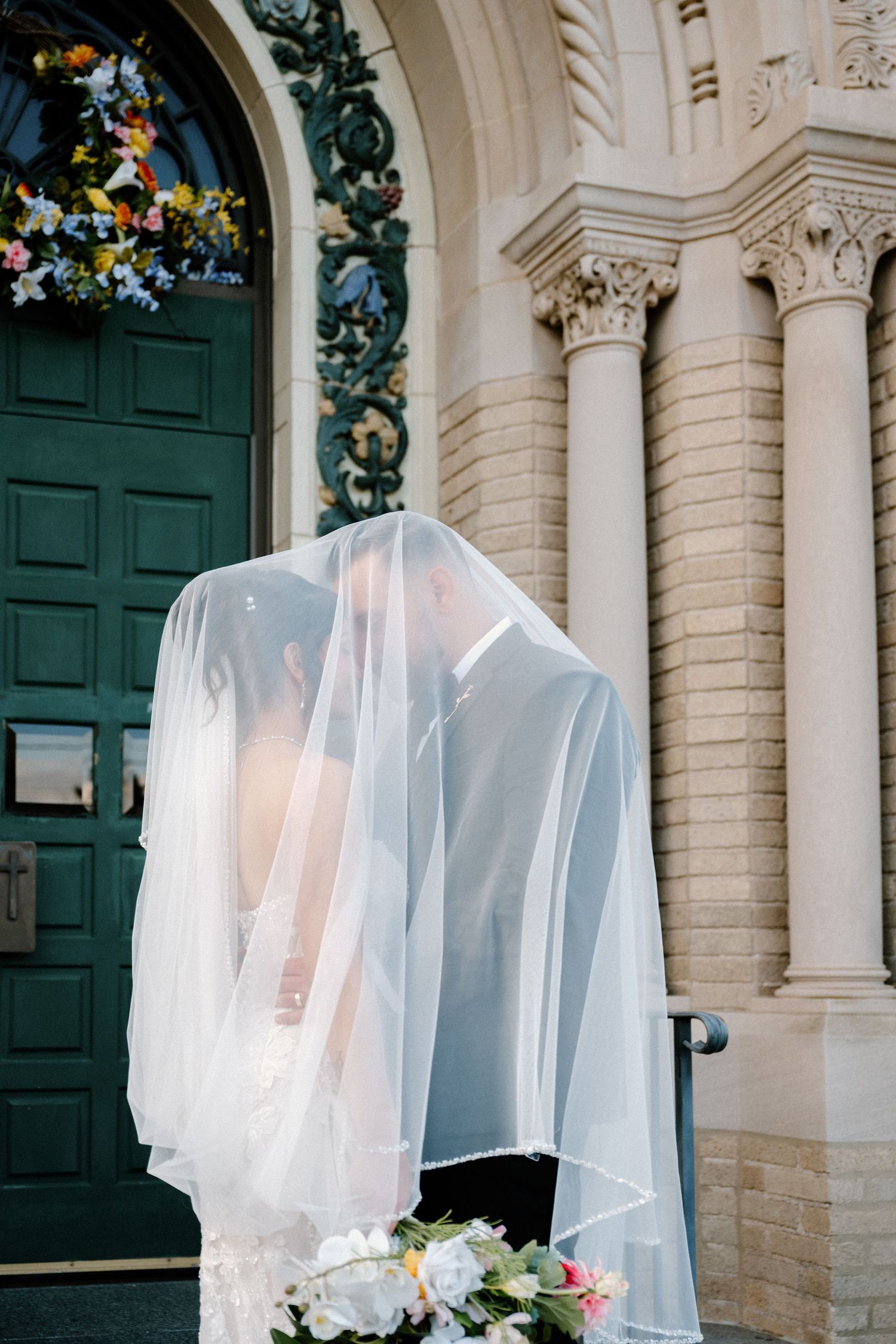 Bride and groom sharing a kiss under a white veil outside an ornate church with green doors and floral arrangements.