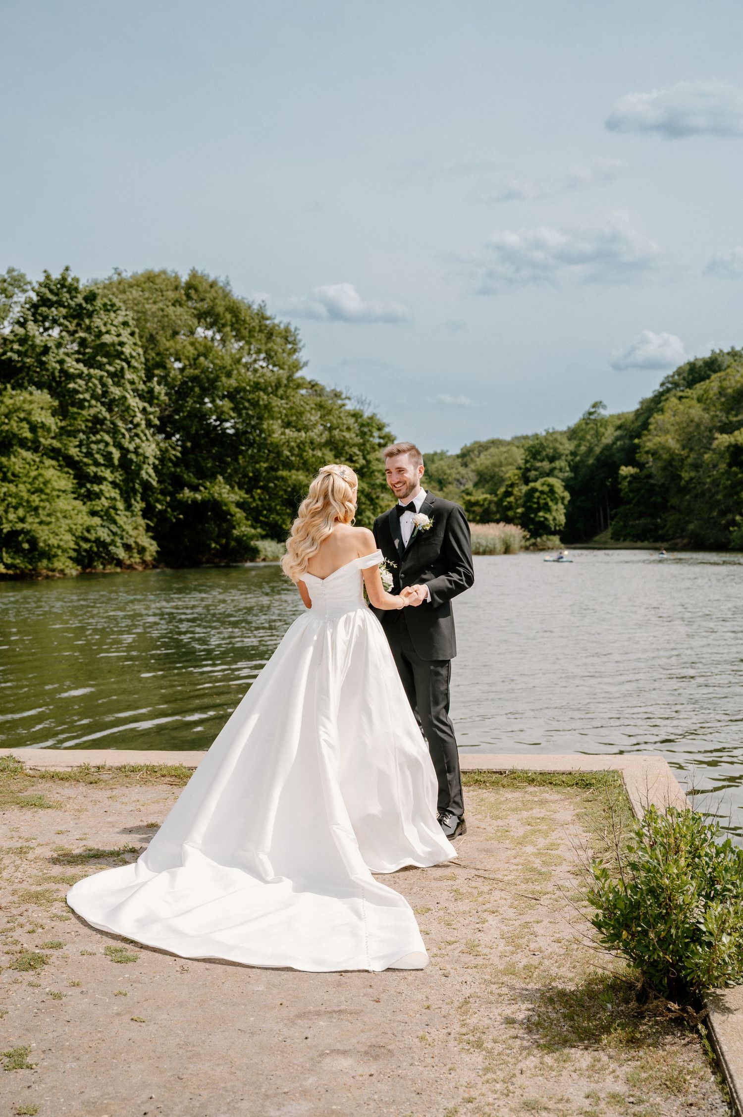 Bride in white off-shoulder gown and groom in black tuxedo pose by a serene lake surrounded by lush green trees.