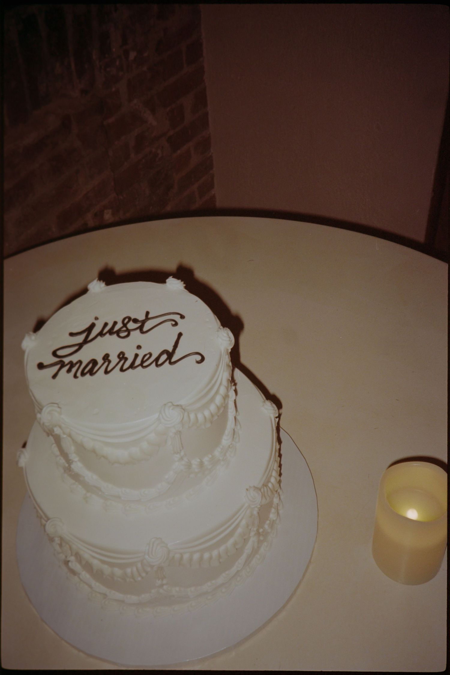 Two-tier white wedding cake with just married inscription on a round table beside a candle against brick wall.