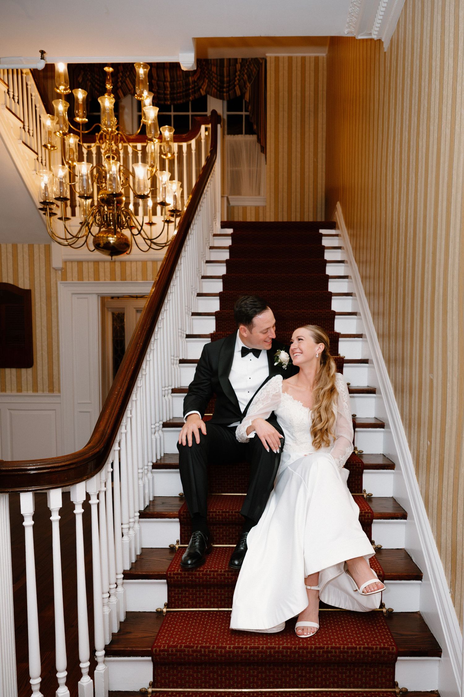 A couple sits together on an elegant staircase with white spindles and red carpet runner under a brass chandelier.