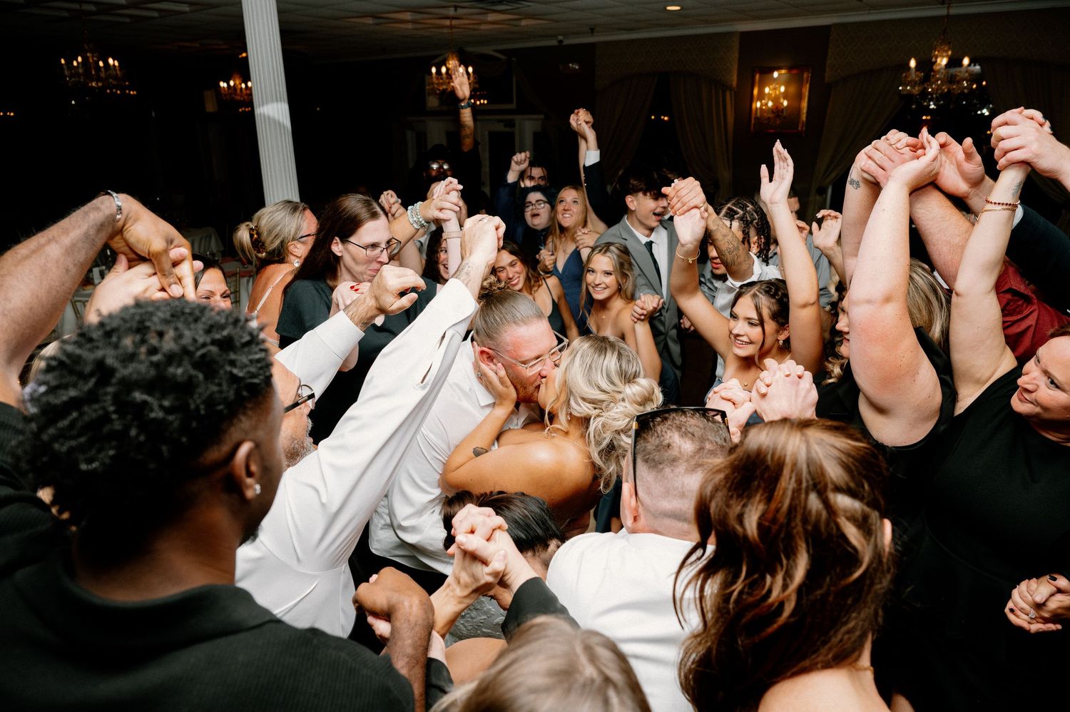 Wedding reception dance floor filled with celebrating guests under dim lighting.