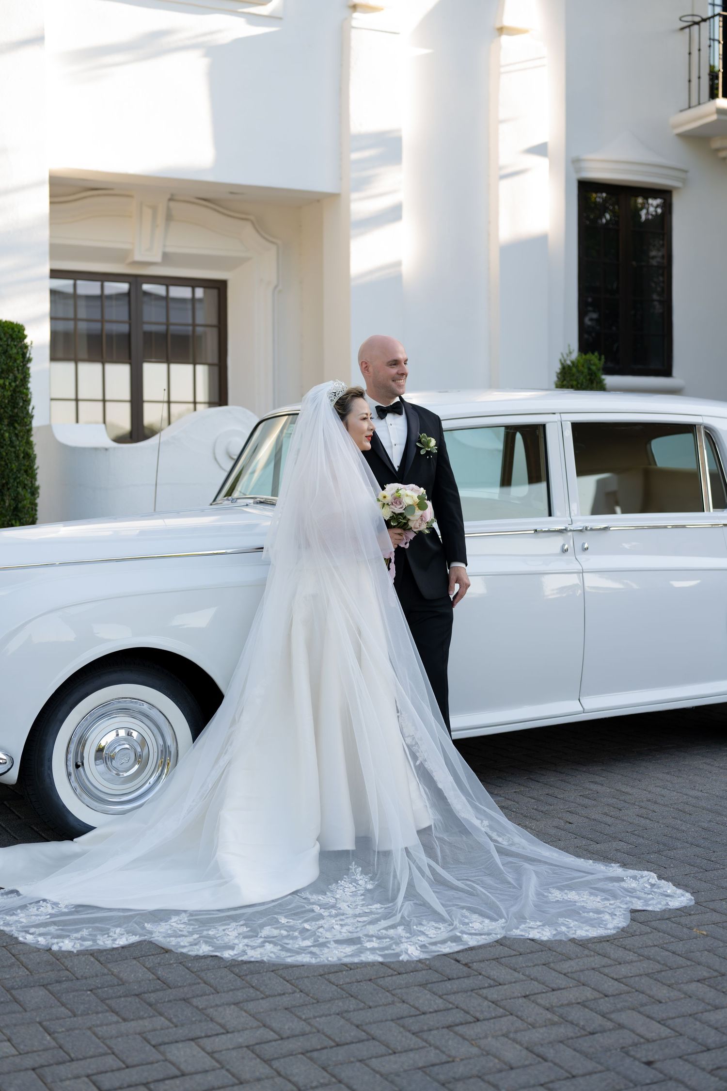 Bride in white gown with long veil and groom in tuxedo pose beside a classic white Rolls-Royce at elegant venue.