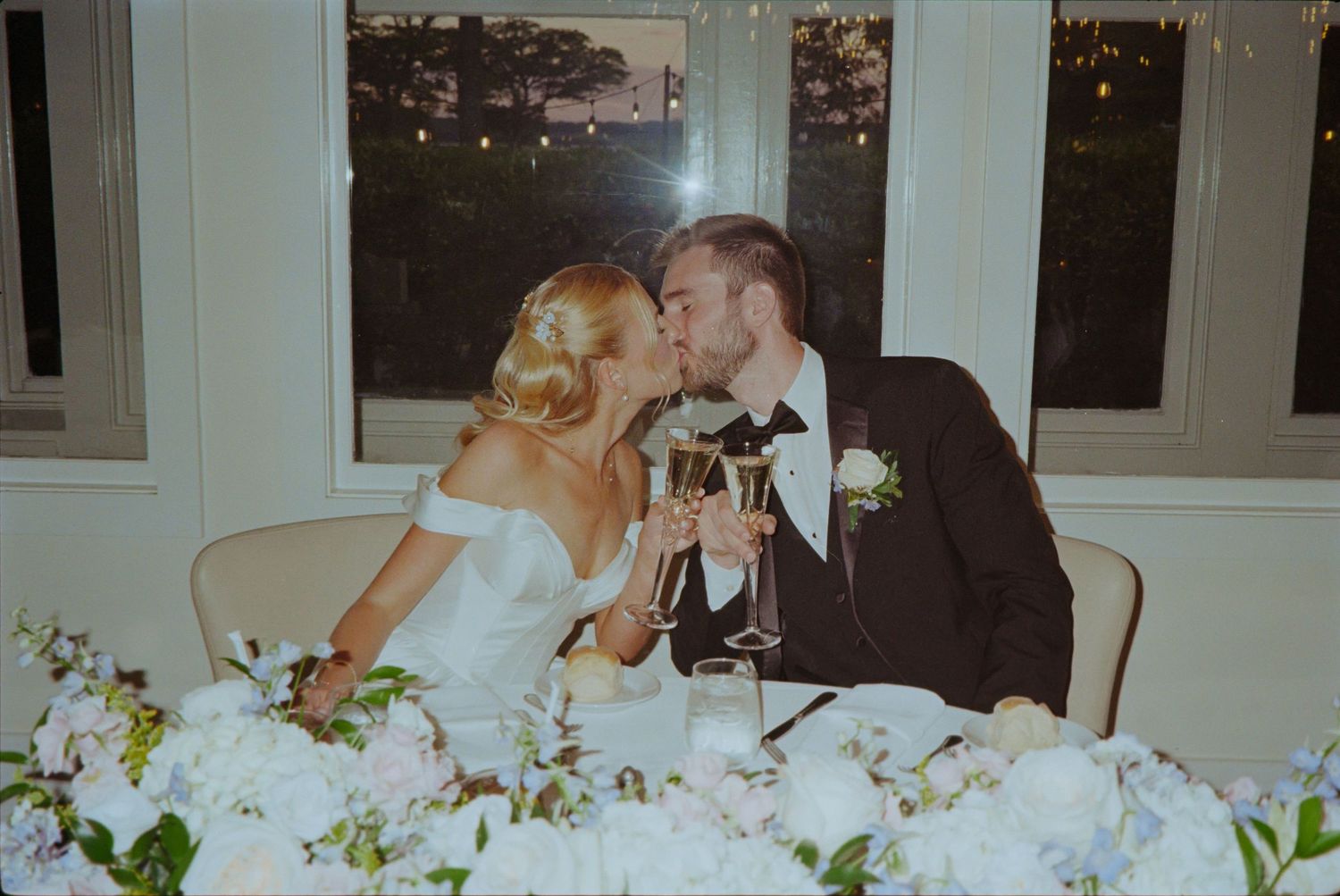 Bride and groom sharing a kiss while clinking champagne glasses at their elegant wedding reception dinner table.