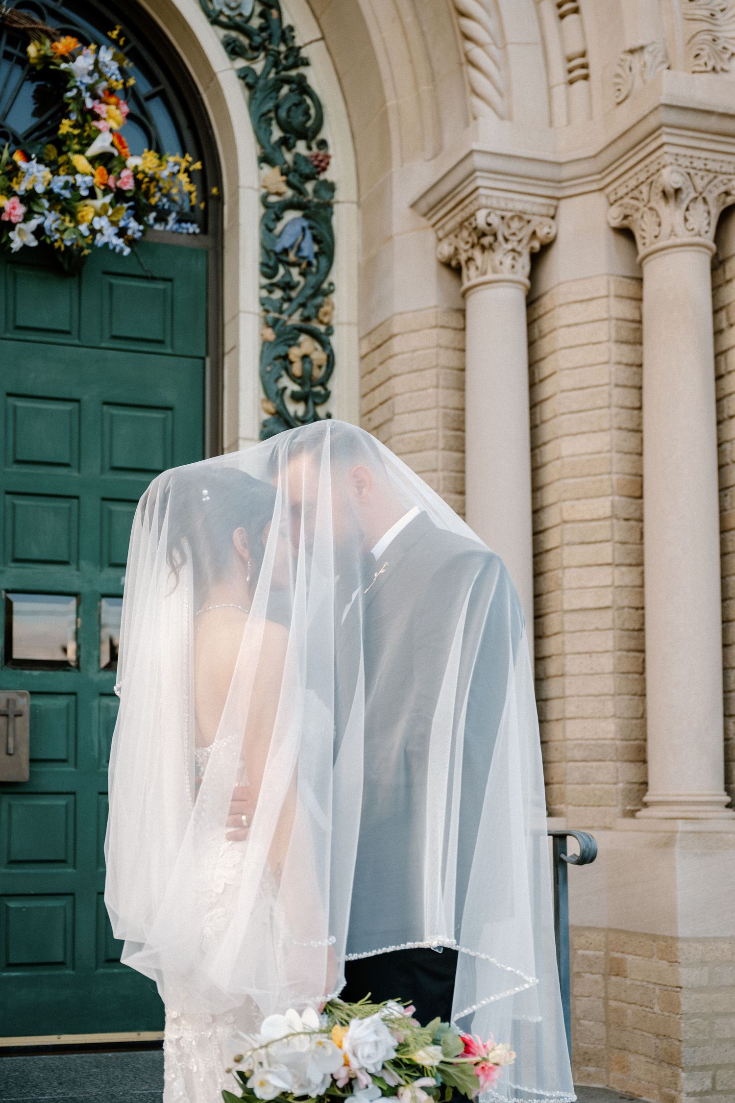 Bride and groom sharing a kiss under a flowing veil outside an ornate church with a green door.