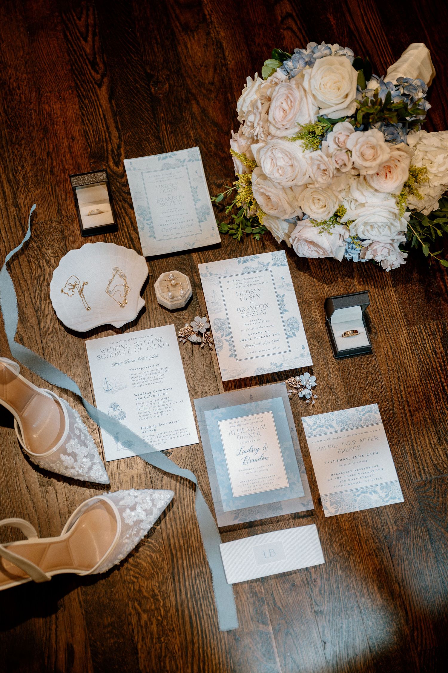 Elegant wedding flat lay featuring dusty blue stationery suite, white floral bouquet, lace heels, and gold rings on wood floor.