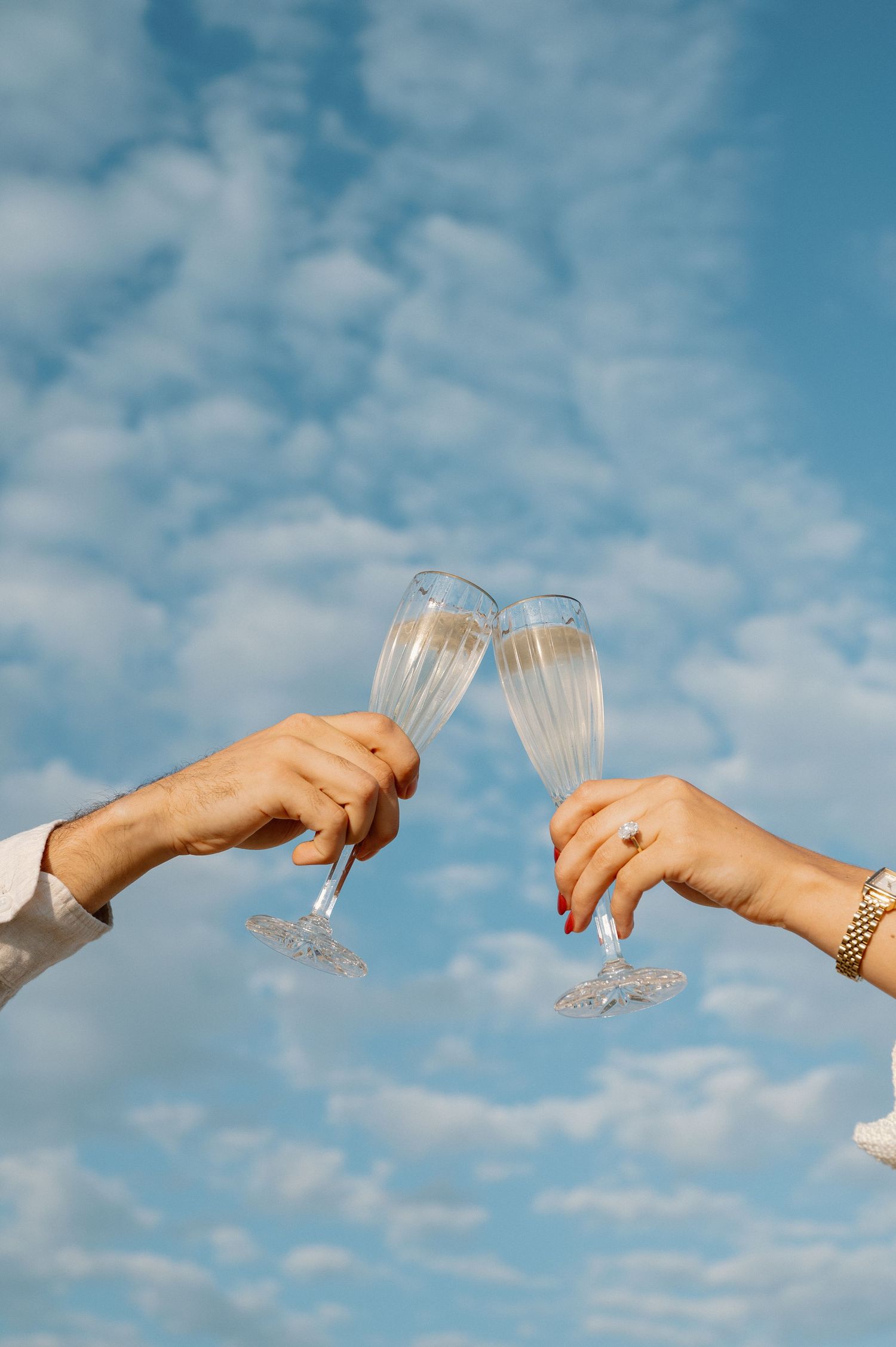Two hands toasting champagne glasses against a blue cloudy sky.