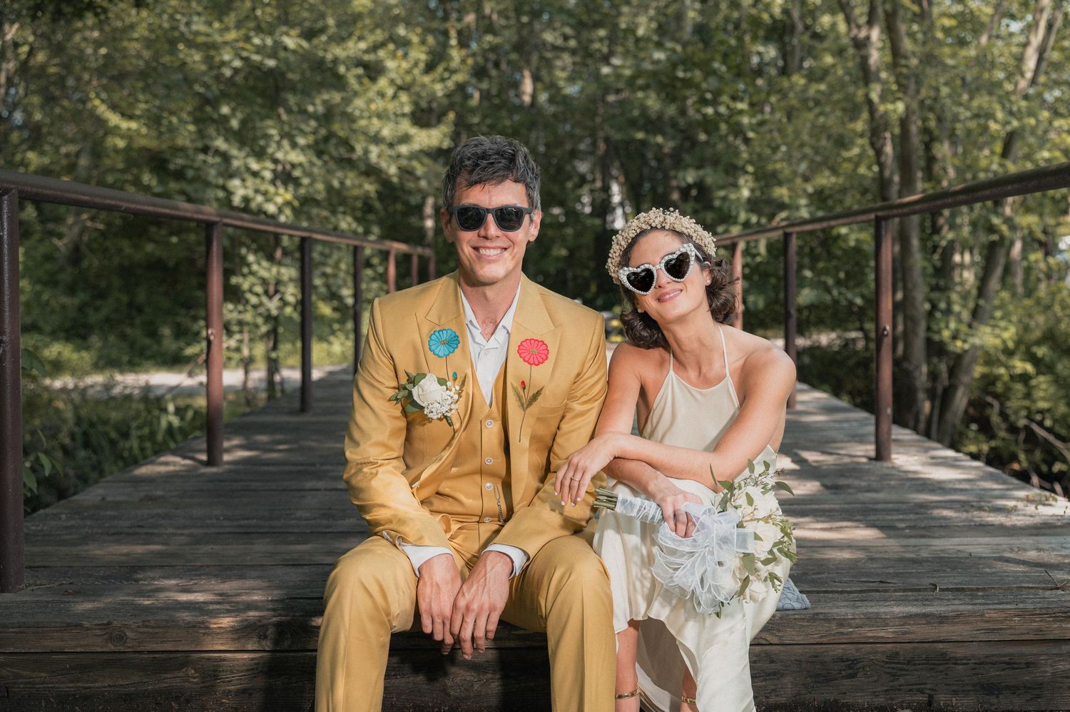 A couple in sunglasses poses on a wooden bridge in a forest setting, with one wearing a yellow jacket.
