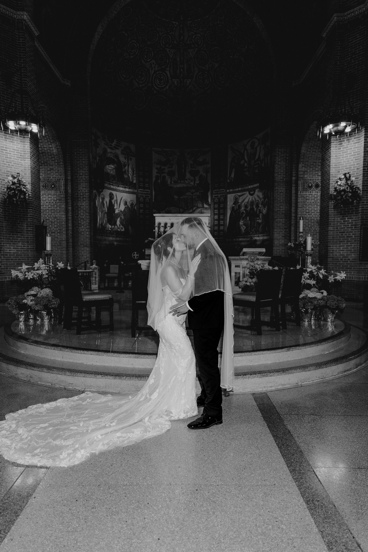 Black and white photo of bride and groom kissing at church altar, long veil trailing on floor.
