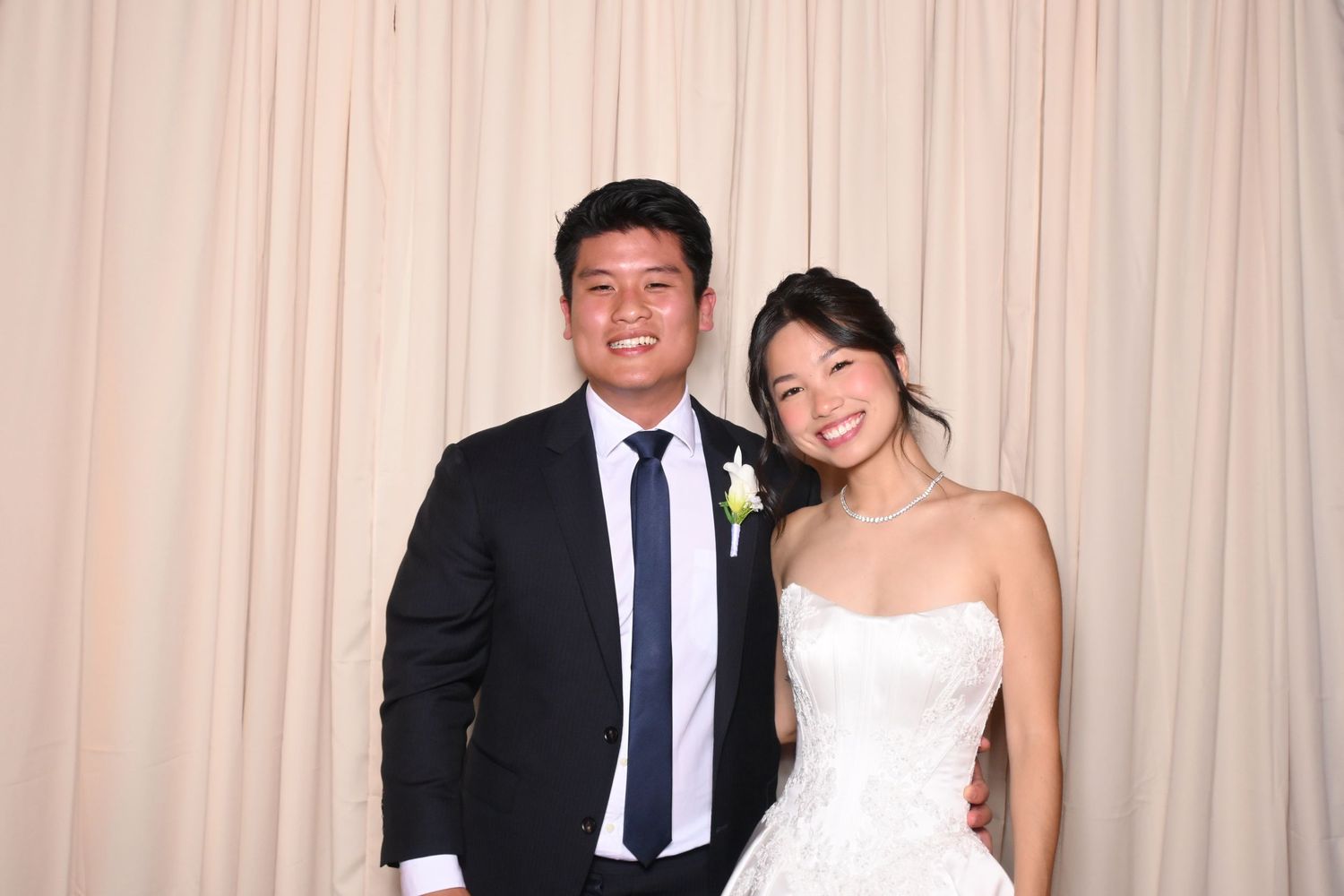 Bride and groom posing together in front of cream backdrop, wearing wedding attire with floral boutonniere.