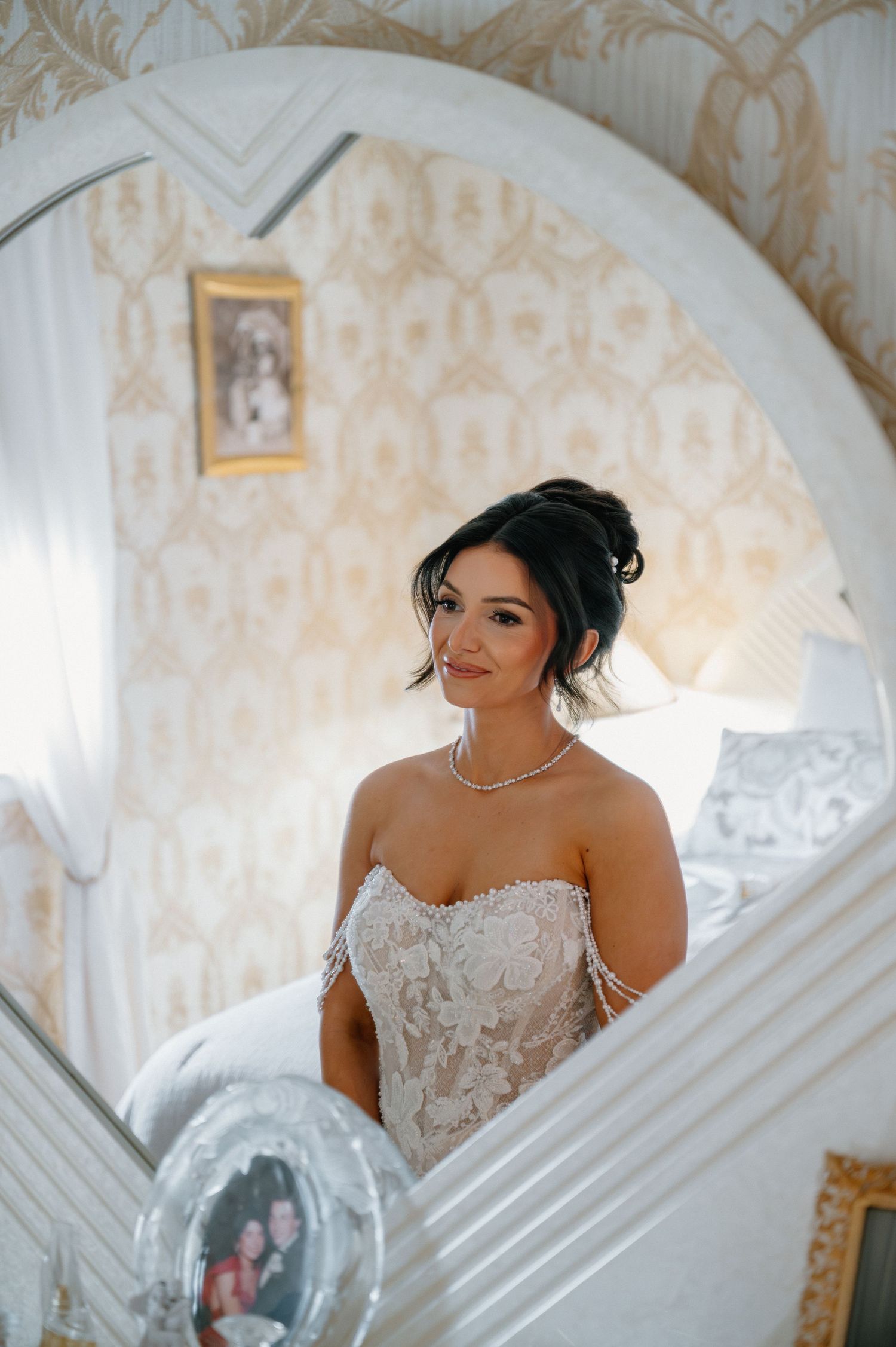 Bride in strapless lace wedding gown with pearl necklace poses elegantly in ornate gold-wallpapered room near arched mirror.