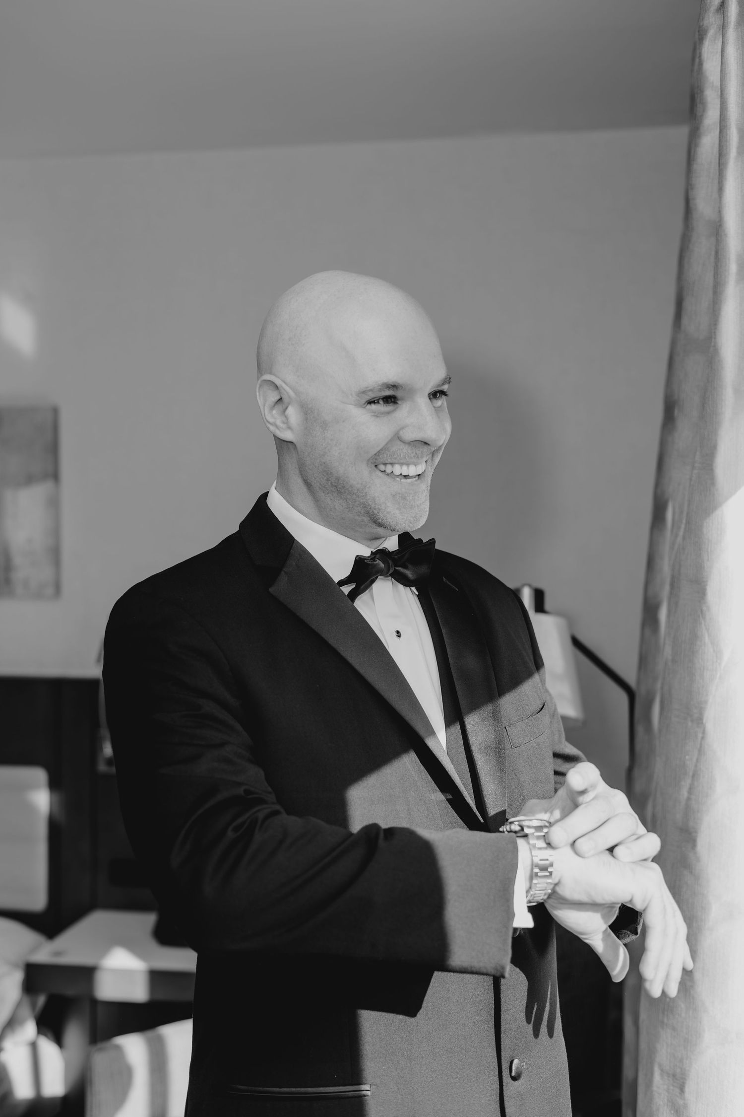 Smiling bald groom in black tuxedo and bow tie adjusting cufflinks in black and white wedding photo.