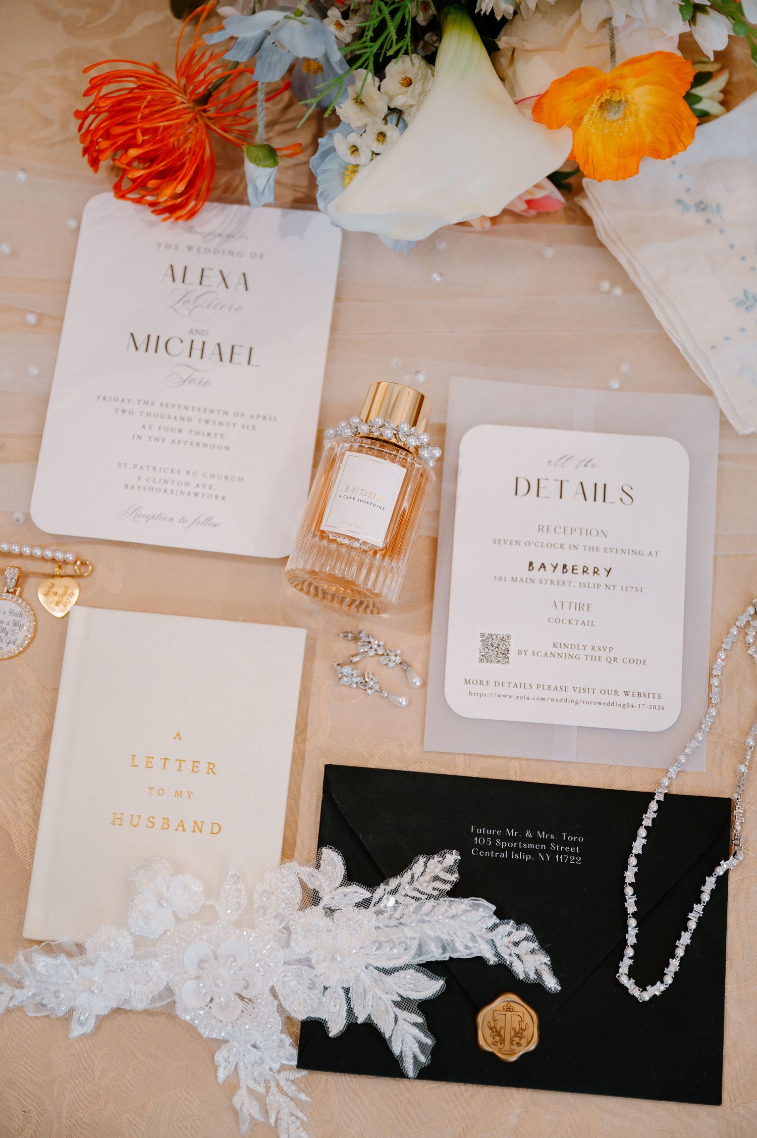 Elegant wedding stationery flatlay featuring invitations, details card, perfume, lace, and jewelry with orange floral accents.