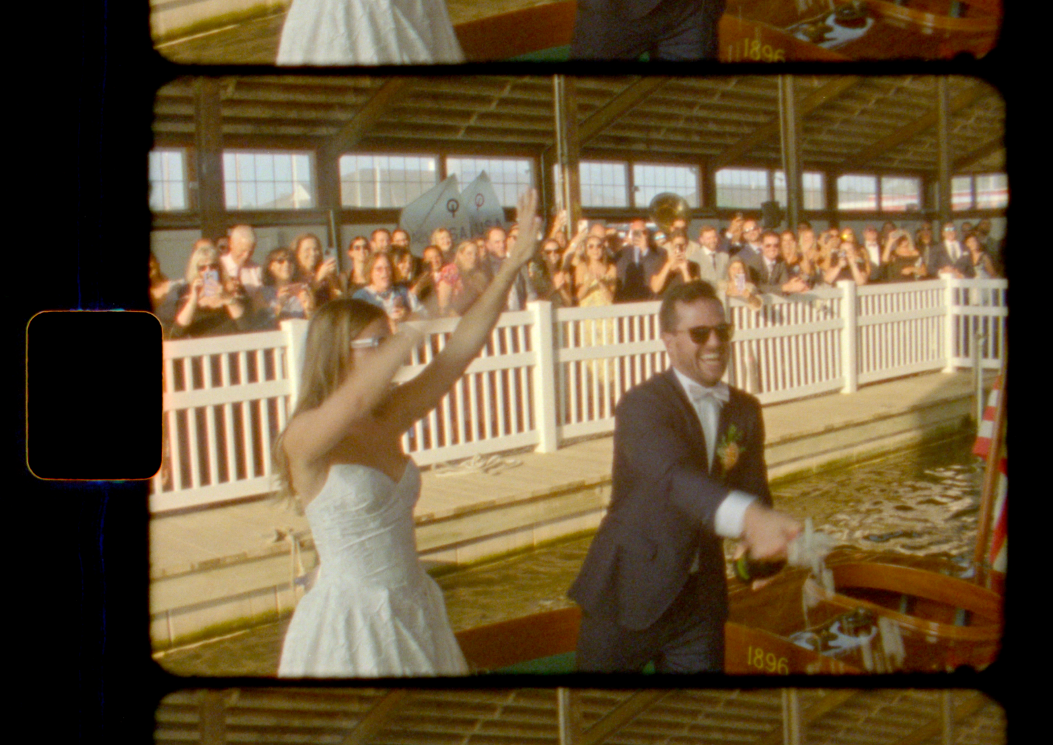 A wedding couple waves to guests from behind a white picket fence during their outdoor celebration.