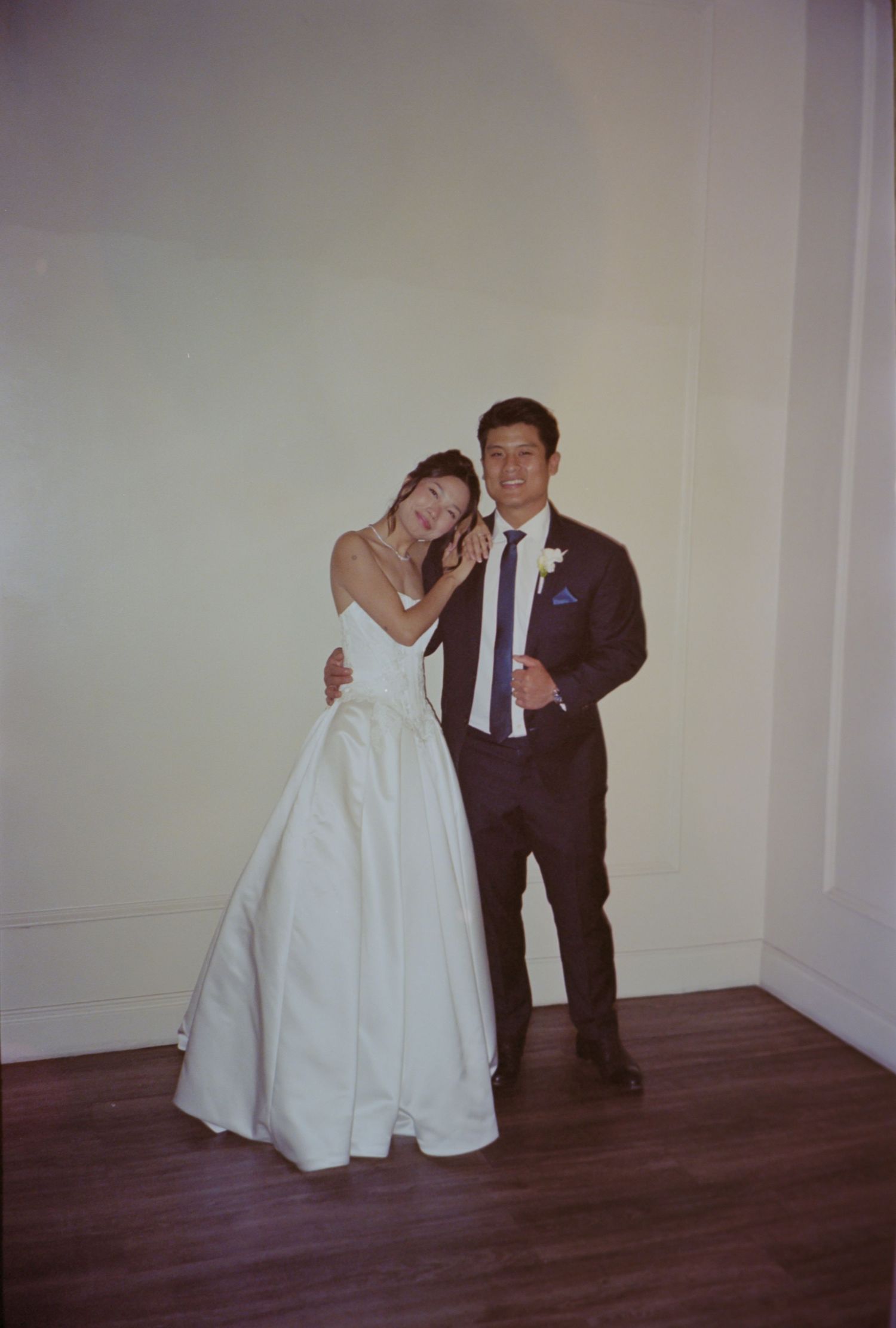Bride in white ball gown leans on groom in dark suit with boutonniere, posing together at wedding reception.