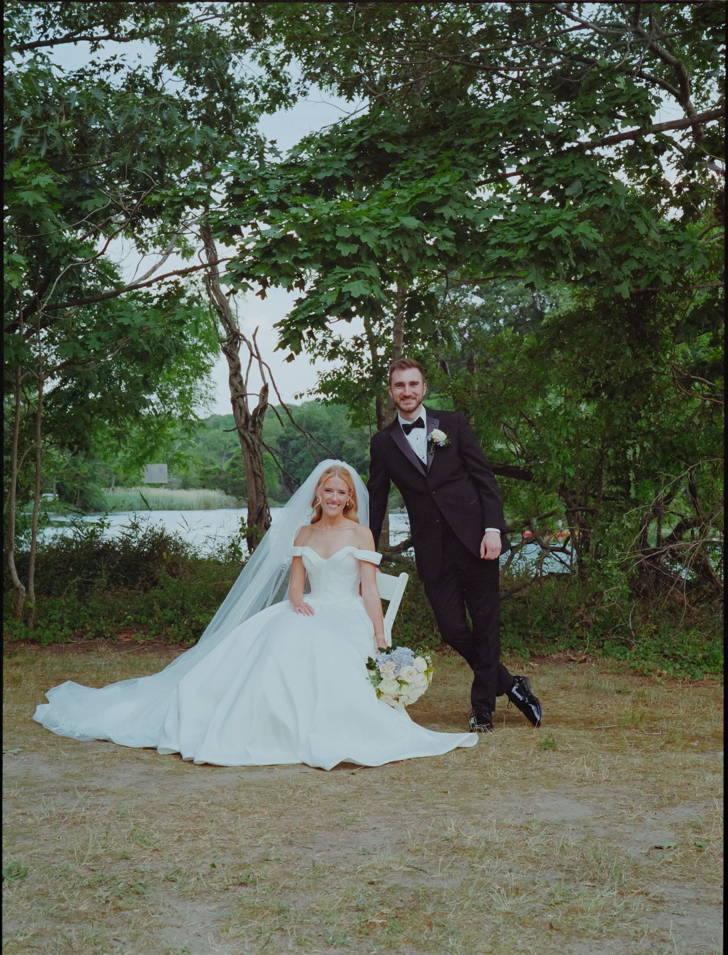 Bride in white off-shoulder gown with veil sits beside groom in black tuxedo outdoors near trees and water.