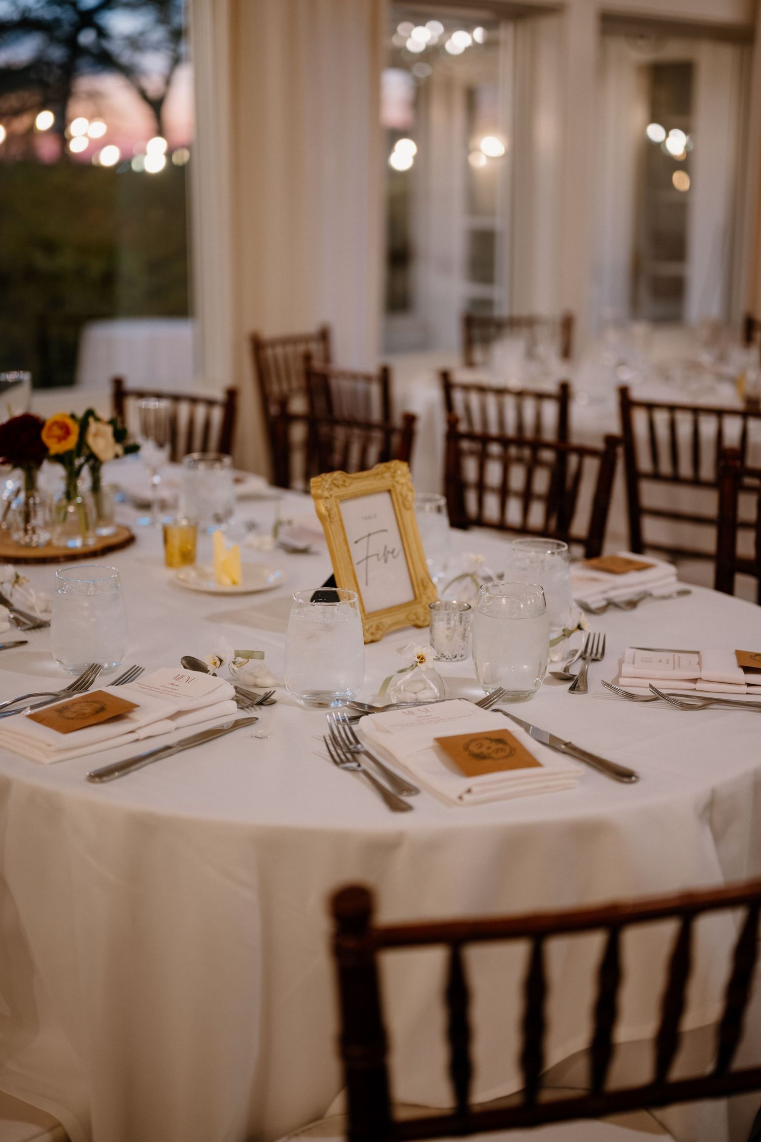 Elegant wedding reception table with white linen, gold framed table number, dark wood chiavari chairs, and floral centerpieces.