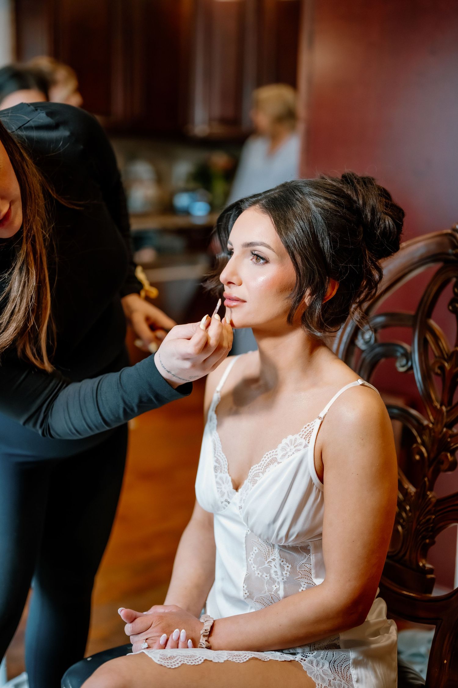 Makeup artist applying lipstick to a bride in a white lace slip dress seated in a wooden chair.