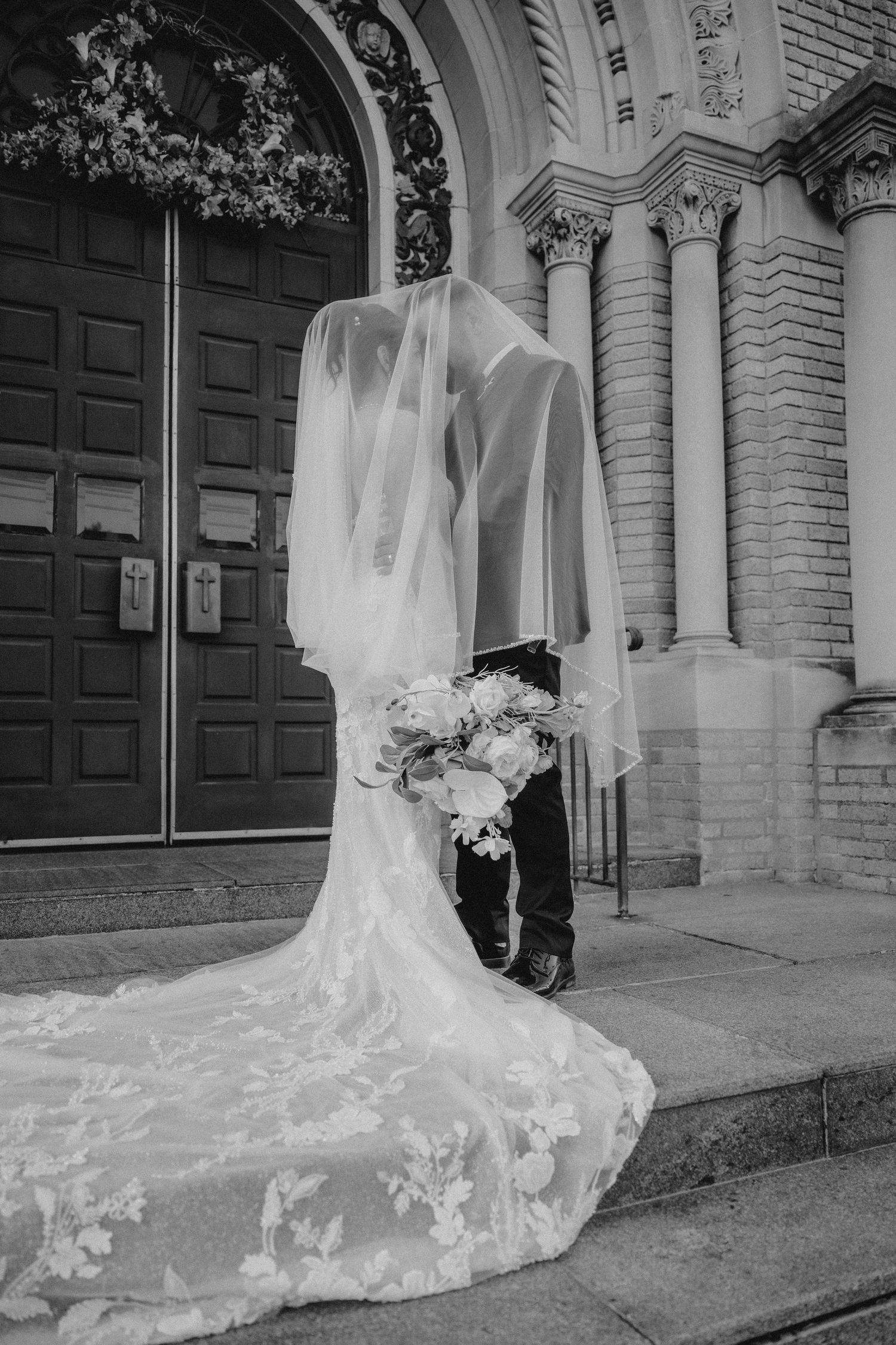 Black and white photo of bride and groom kissing on church steps, veil flowing over them, lace wedding dress train spread out.