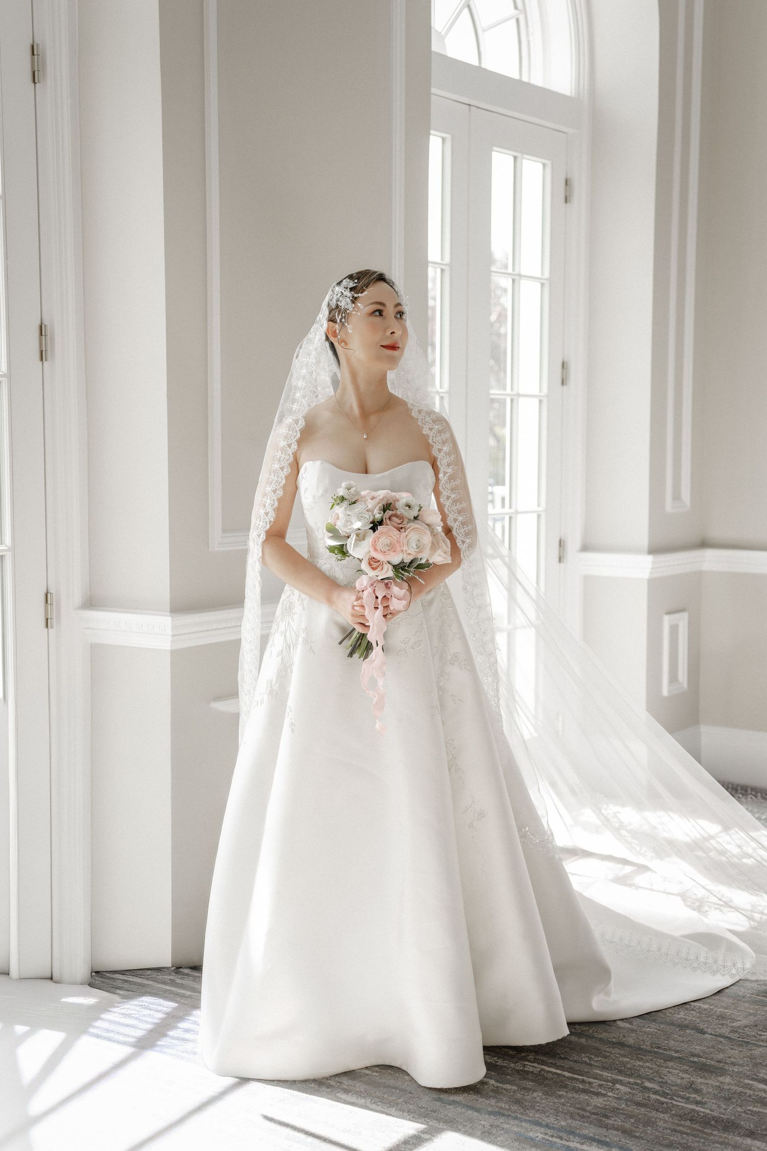 Bride in strapless white ball gown with lace veil holding pink floral bouquet standing by bright windows.