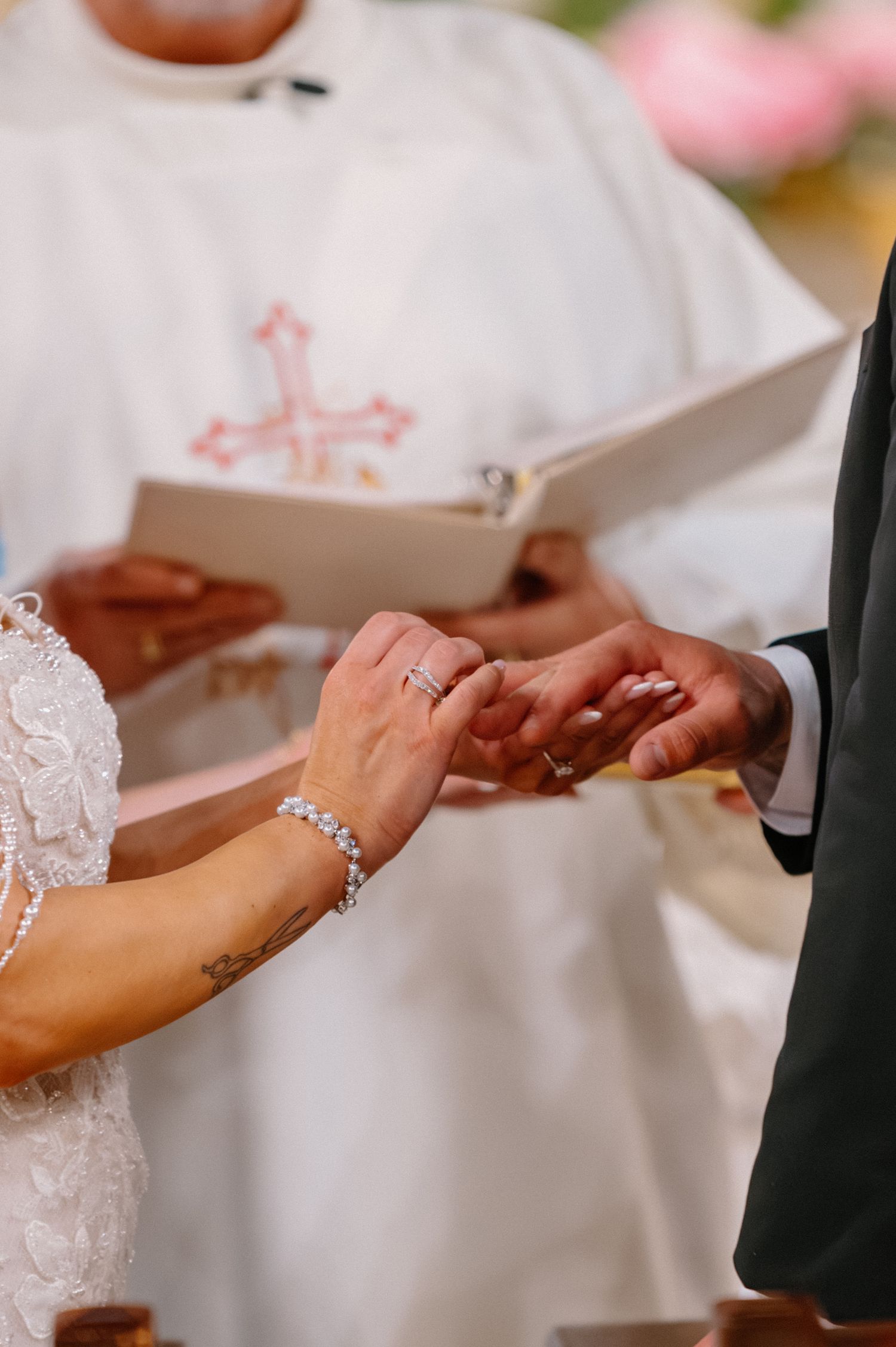 Bride placing a wedding ring on groom's finger during a Christian ceremony, officiant holding a Bible in background.