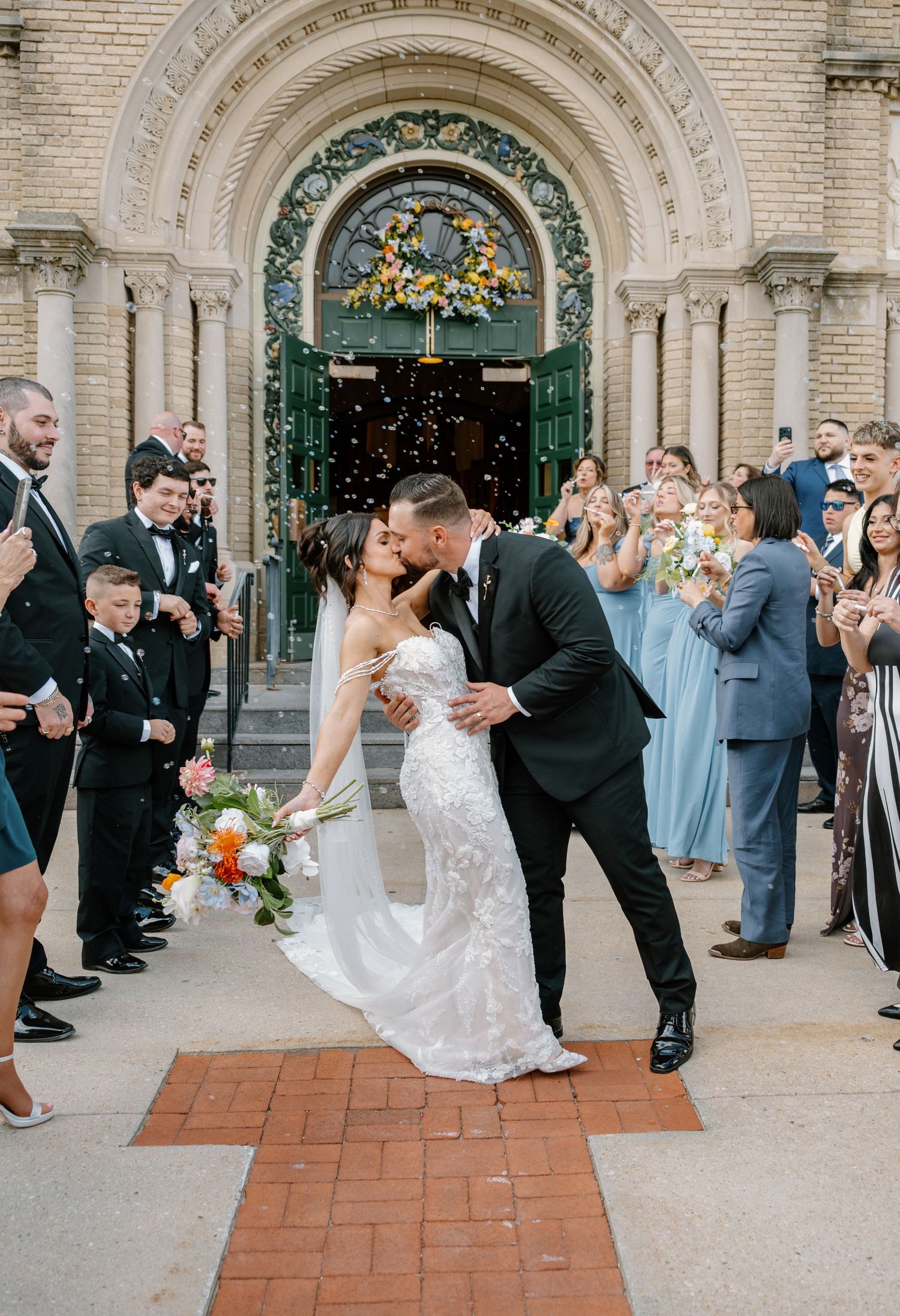 Bride and groom share a romantic kiss outside an ornate church entrance surrounded by cheering wedding guests.