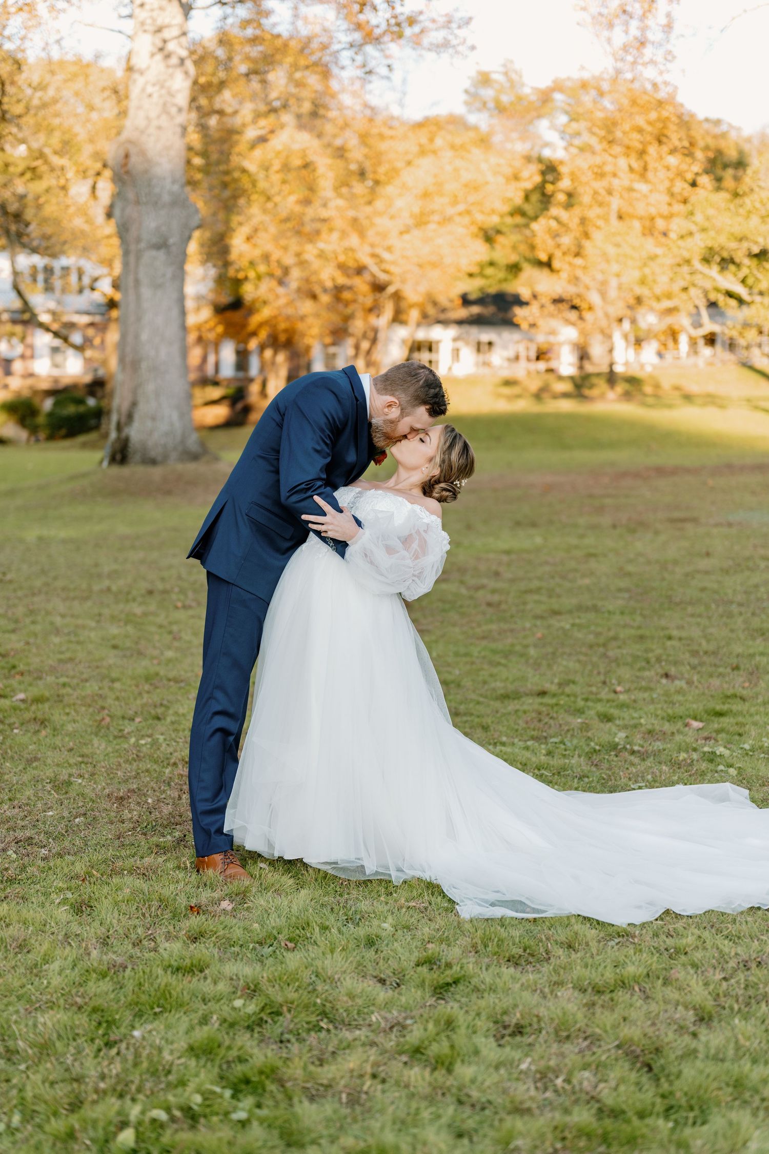 A romantic wedding couple shares a kiss on a grassy lawn surrounded by autumn trees with golden foliage.