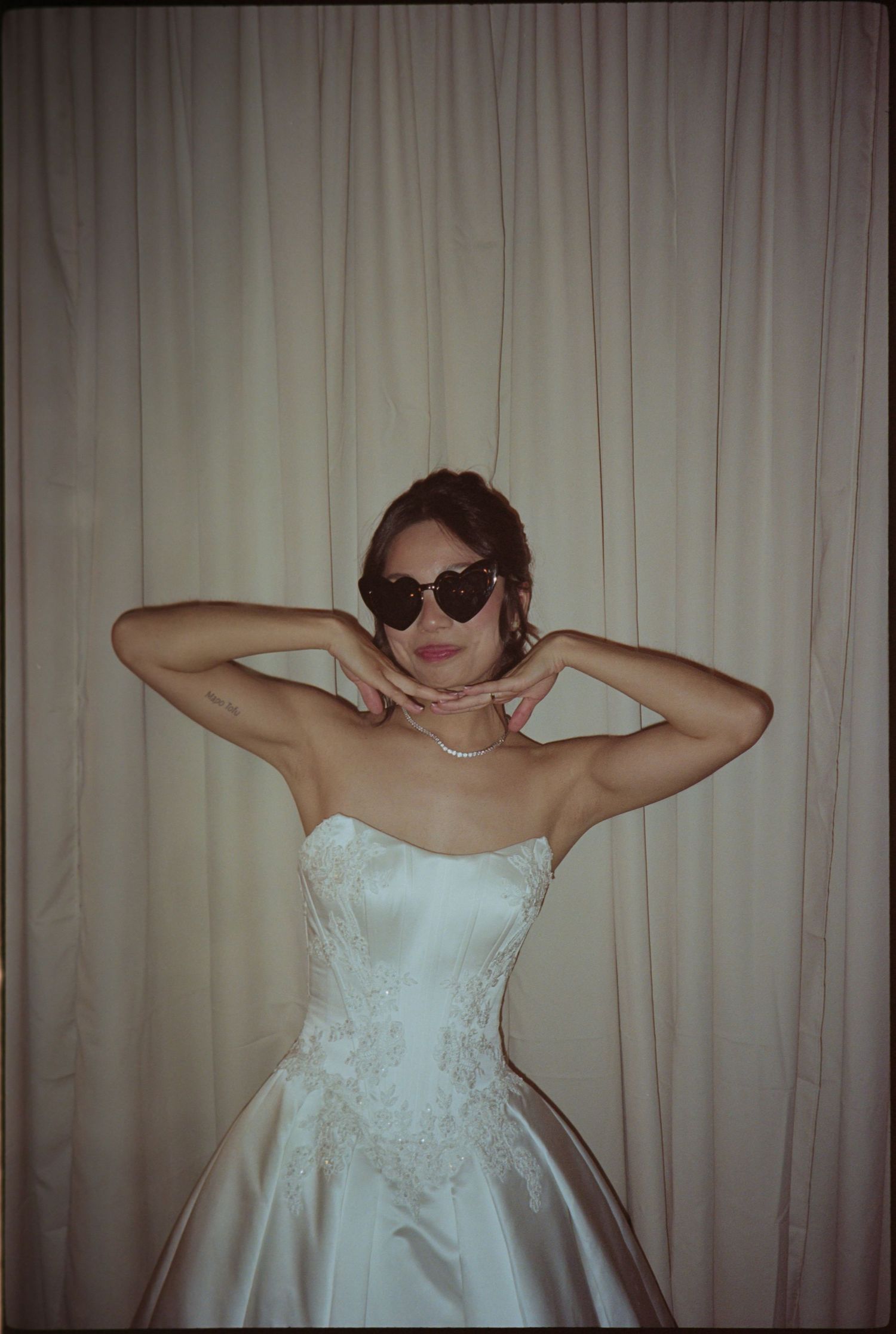 Woman in strapless white wedding gown and heart-shaped sunglasses posing with hands behind head against cream curtain.