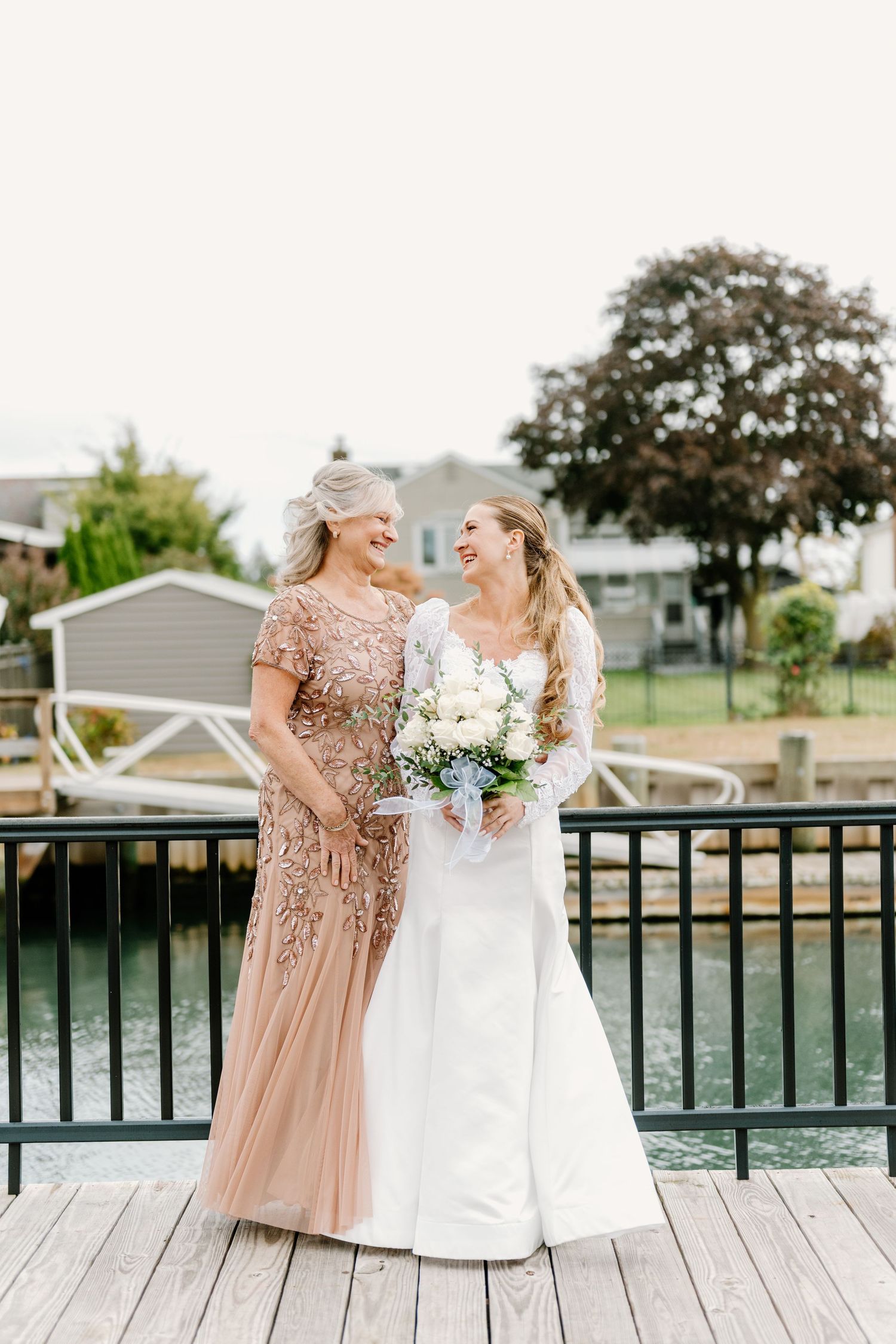Wedding party in rose gold and white dresses laughing together on waterfront deck.