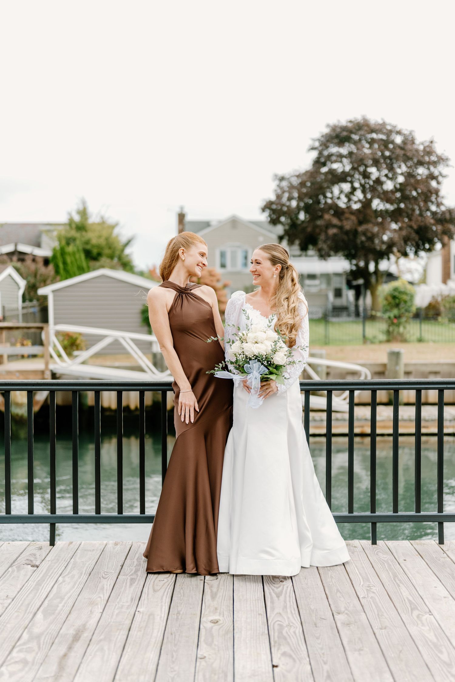 Bride in white gown holds bouquet beside bridesmaid in brown satin dress on waterfront deck.