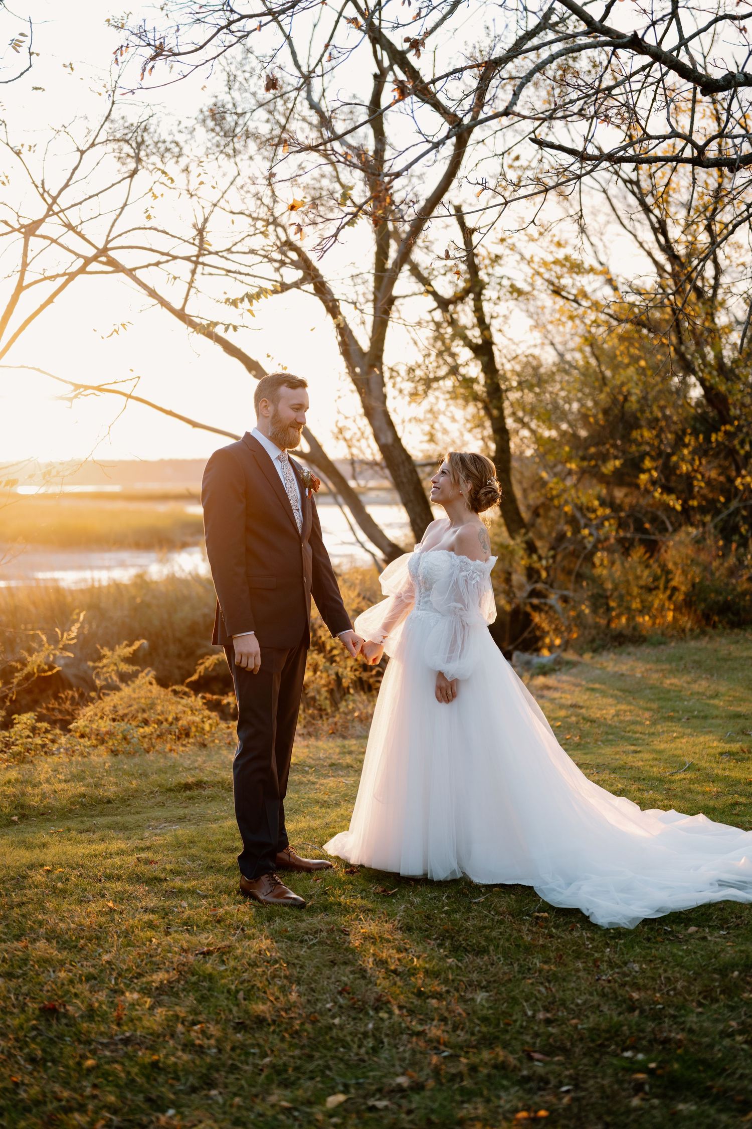 A couple in wedding attire shares an intimate moment at sunset in a scenic outdoor setting with bare trees.