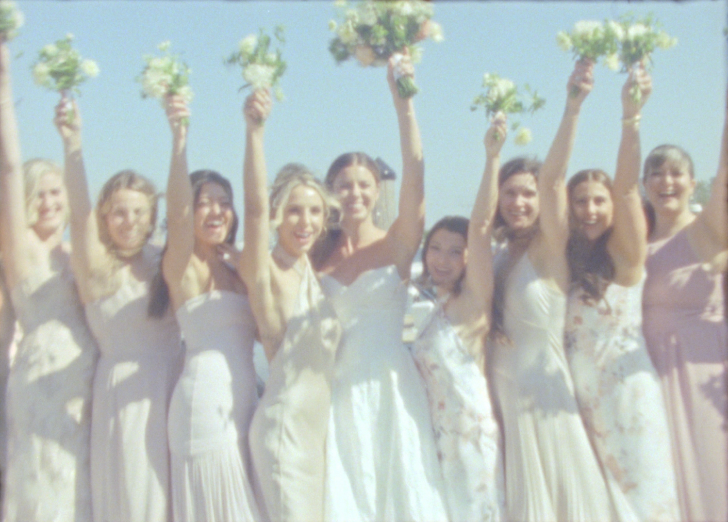 Bridesmaids in light dresses raising bouquets against blue sky.