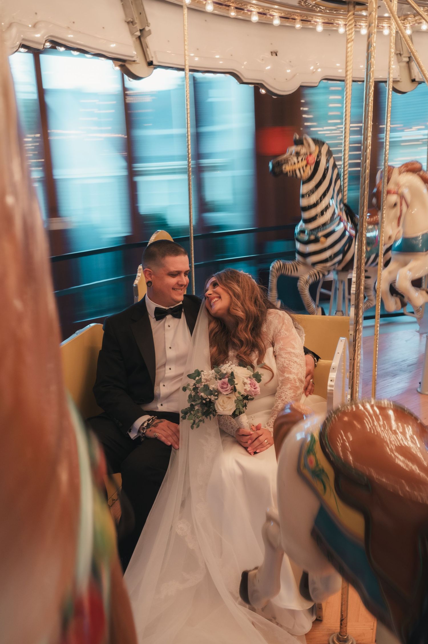 A couple in wedding attire share a romantic moment on a carousel with decorative horses in the background.
