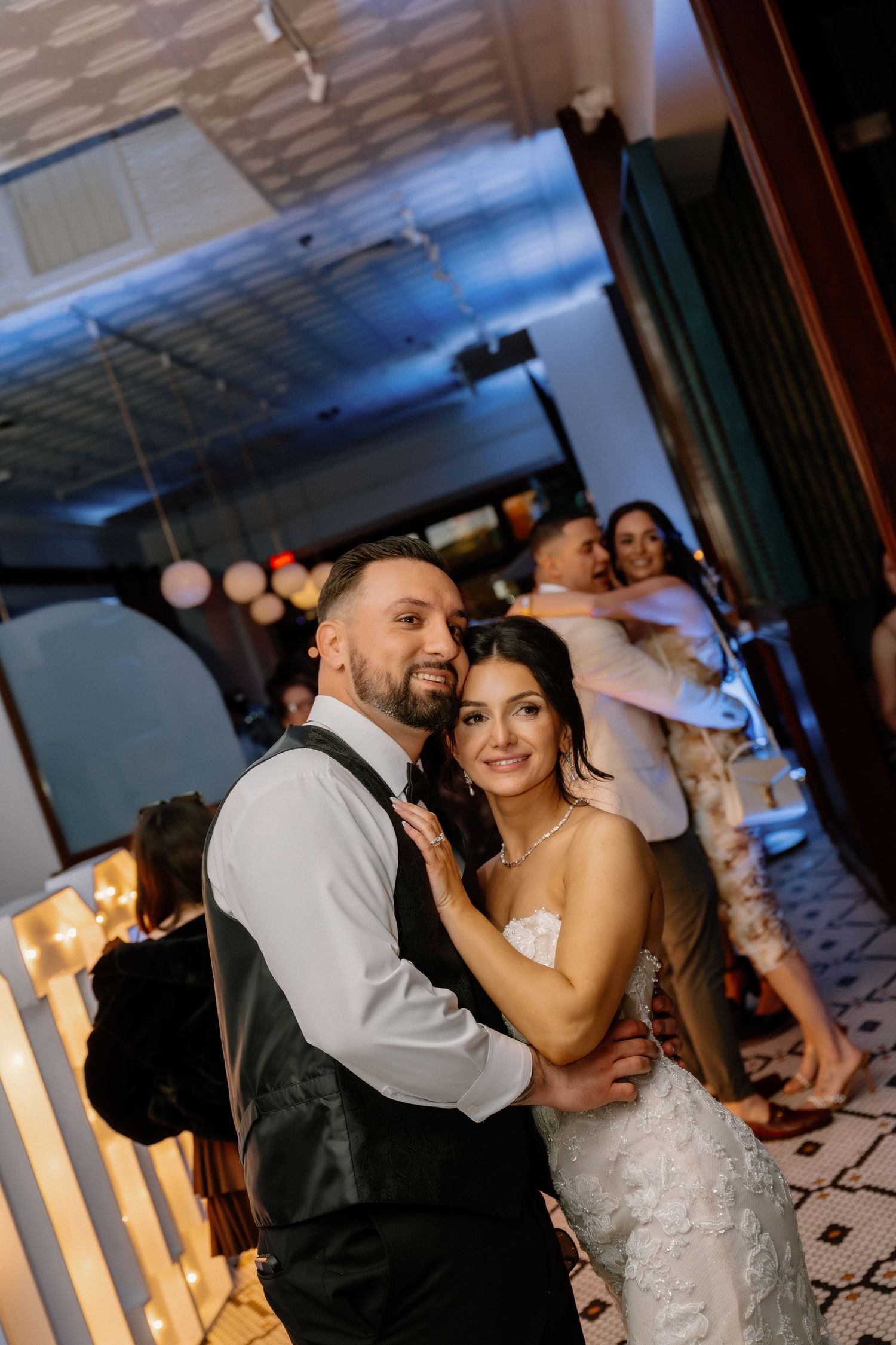 Bride and groom share first dance at wedding reception, smiling at camera with guests dancing in background.
