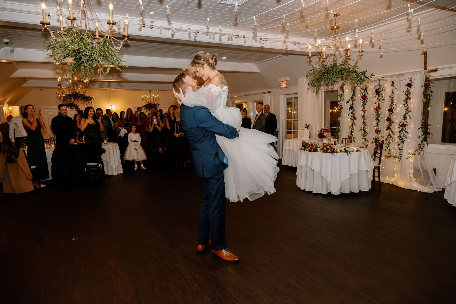 Groom lifts bride during first dance at elegant wedding reception with floral chandeliers and candlelit venue.