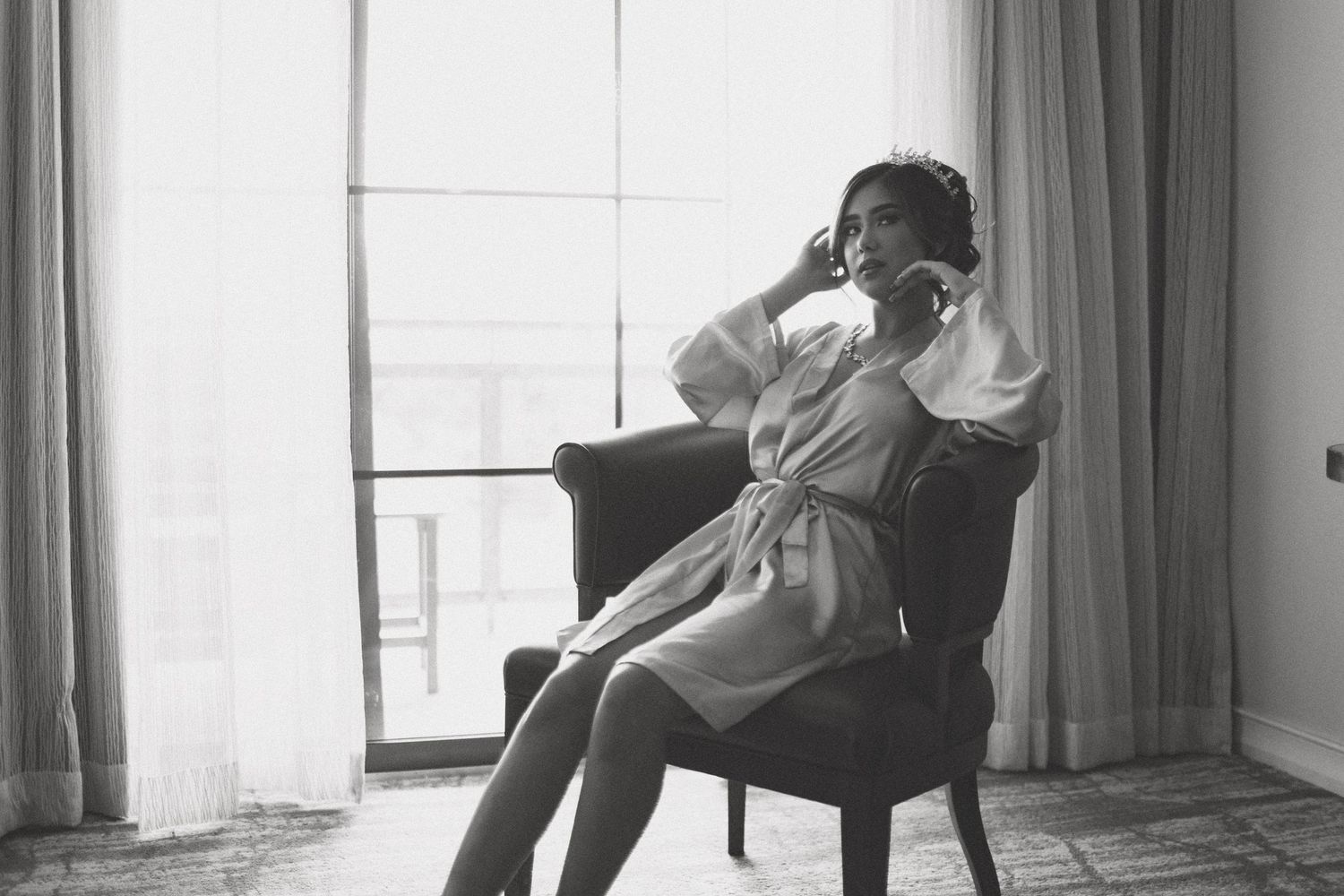 A moody black and white portrait of someone sitting thoughtfully in an armchair by a window.
