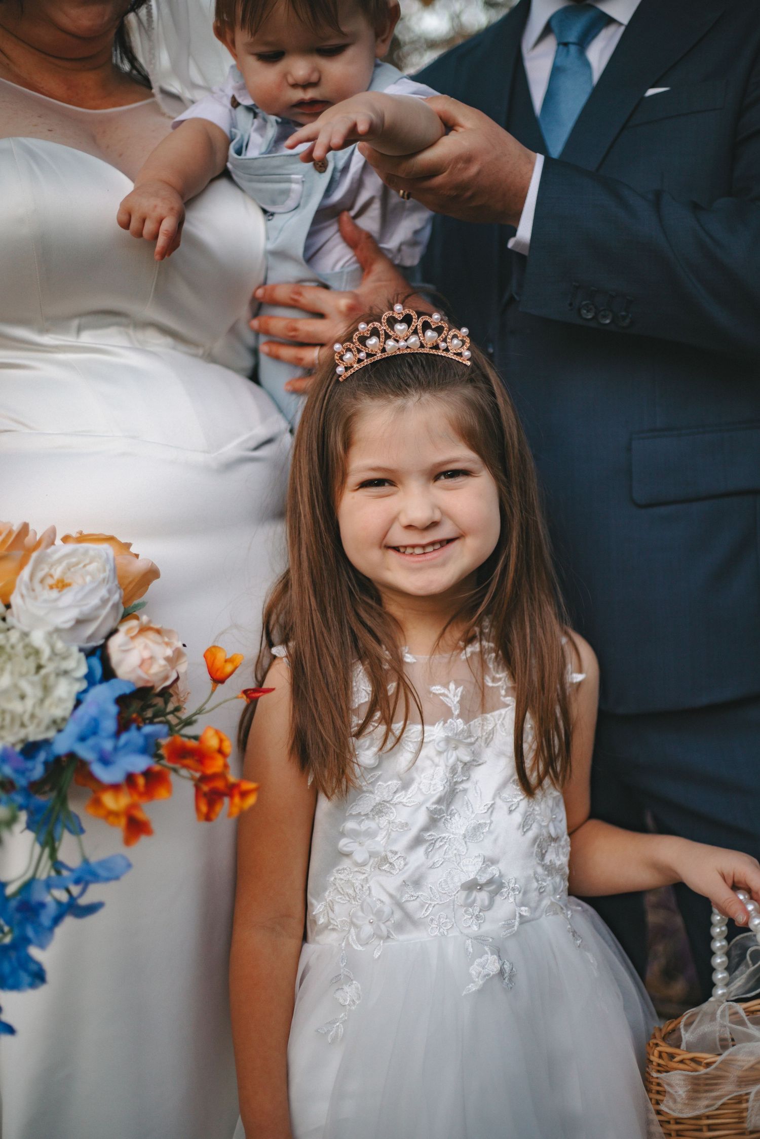Close-up of flower girl in white lace dress smiling at outdoor wedding.