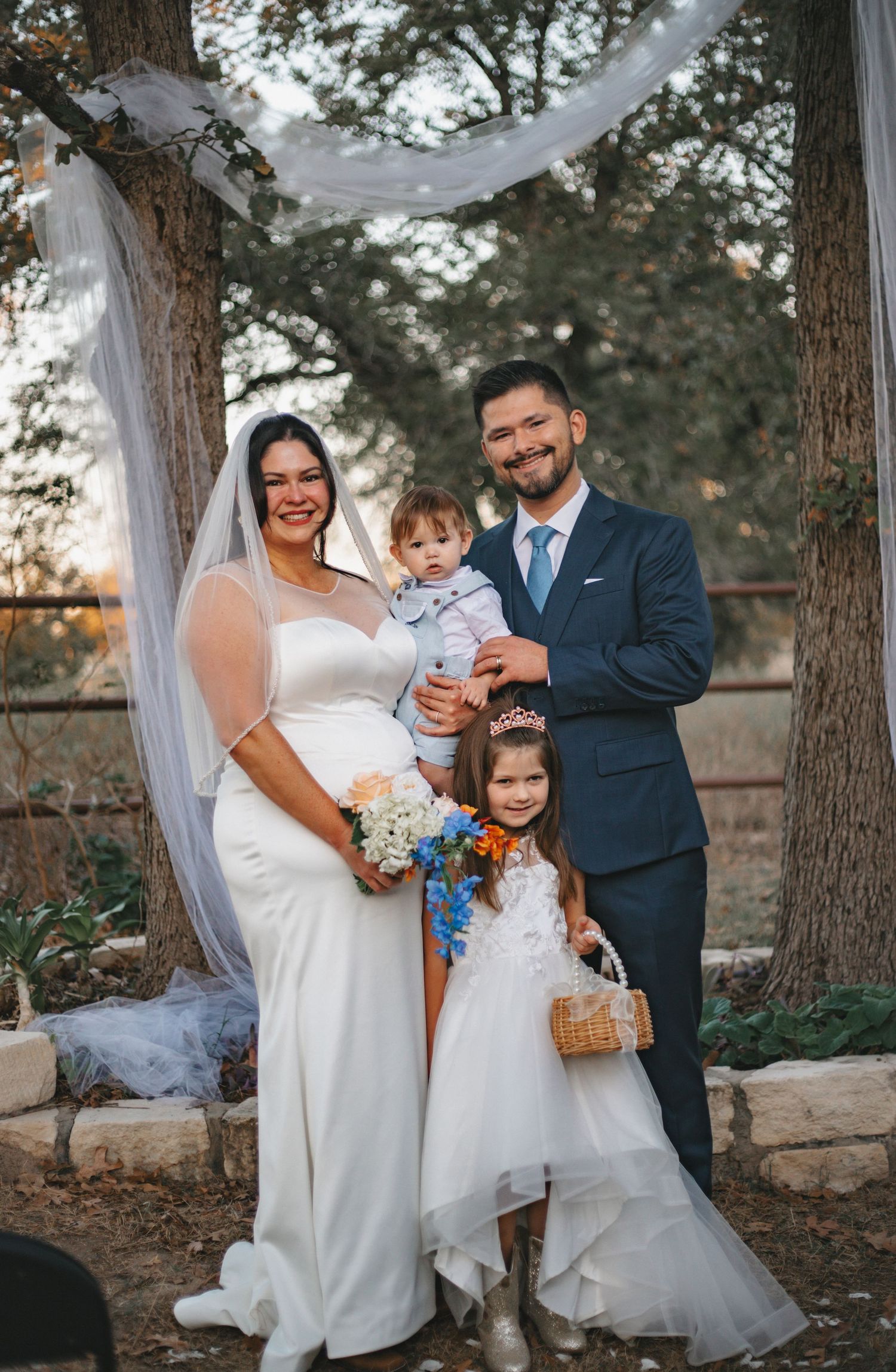 Wedding party poses outdoors with flower girl holding basket under white draping and trees.