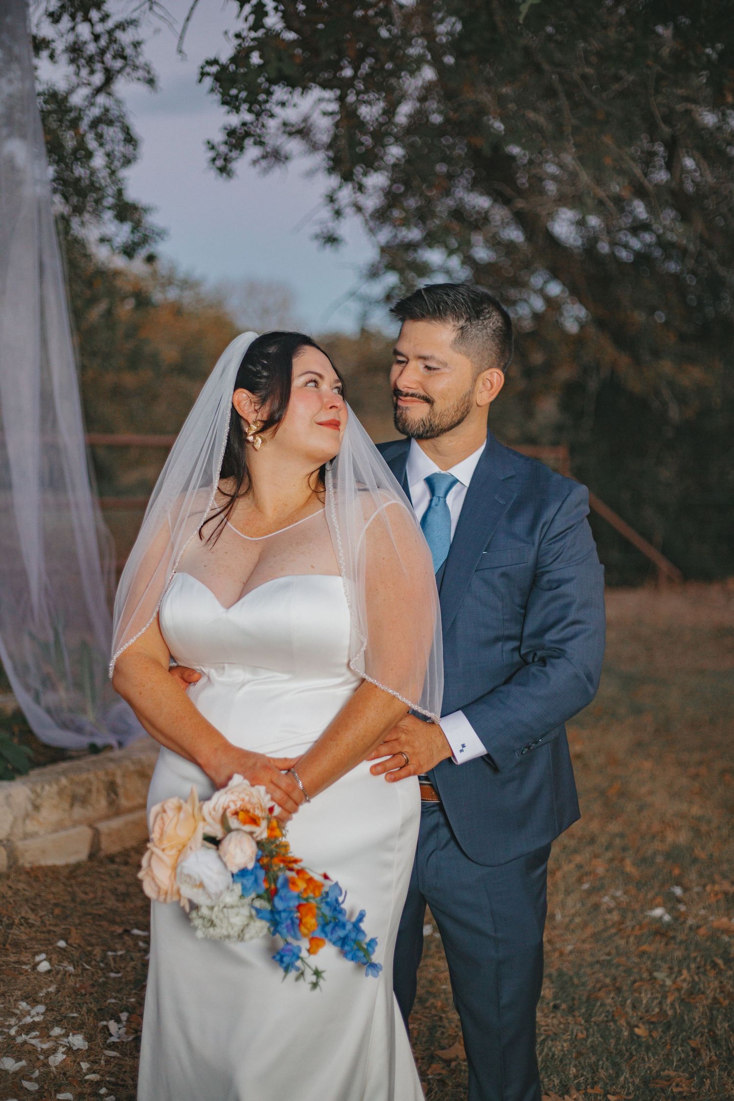 Couple shares intimate moment while holding blue and orange bouquet at outdoor wedding.