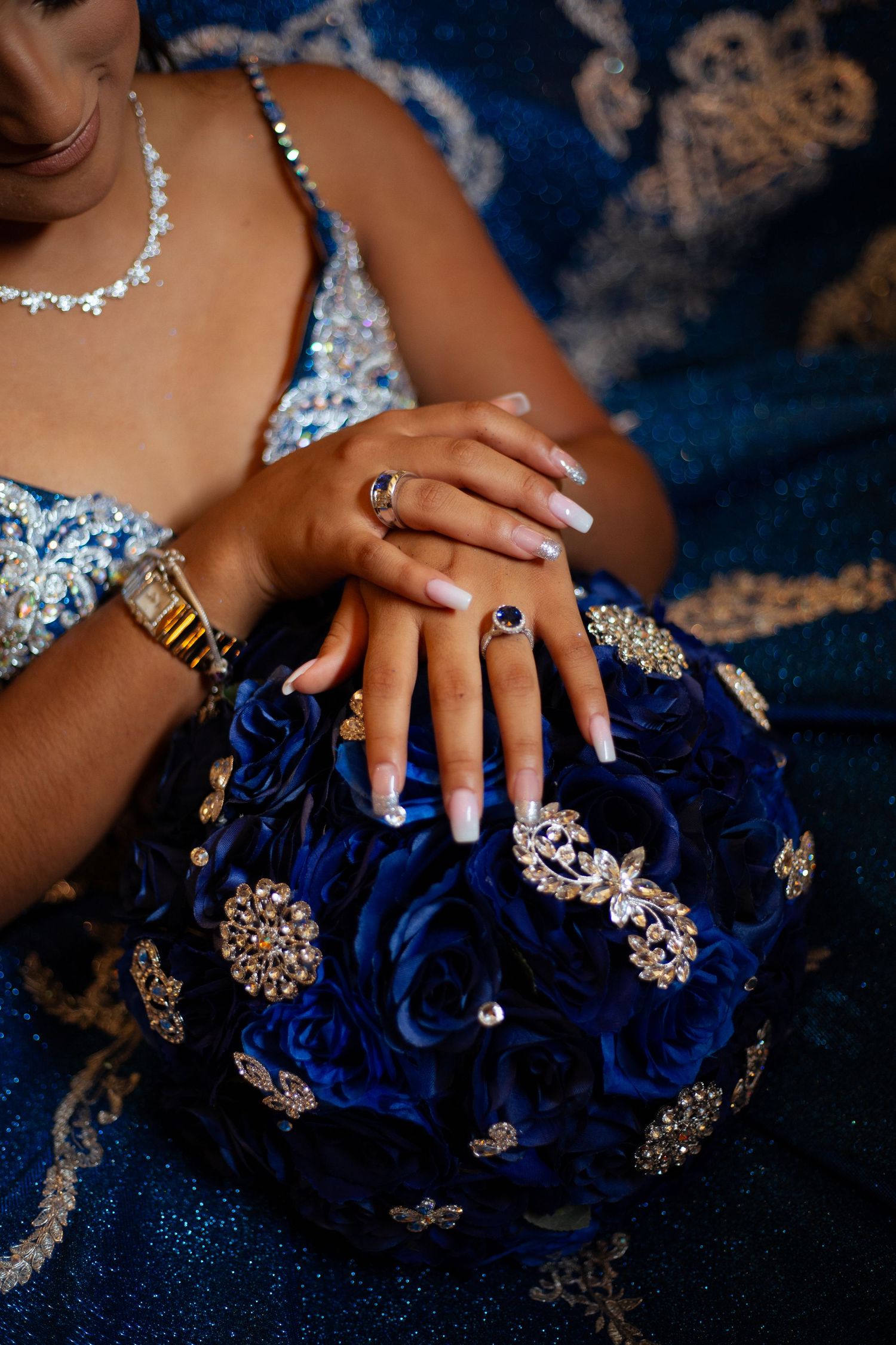 Close-up of manicured hands with jewelry against dark blue fabric with embellishments.