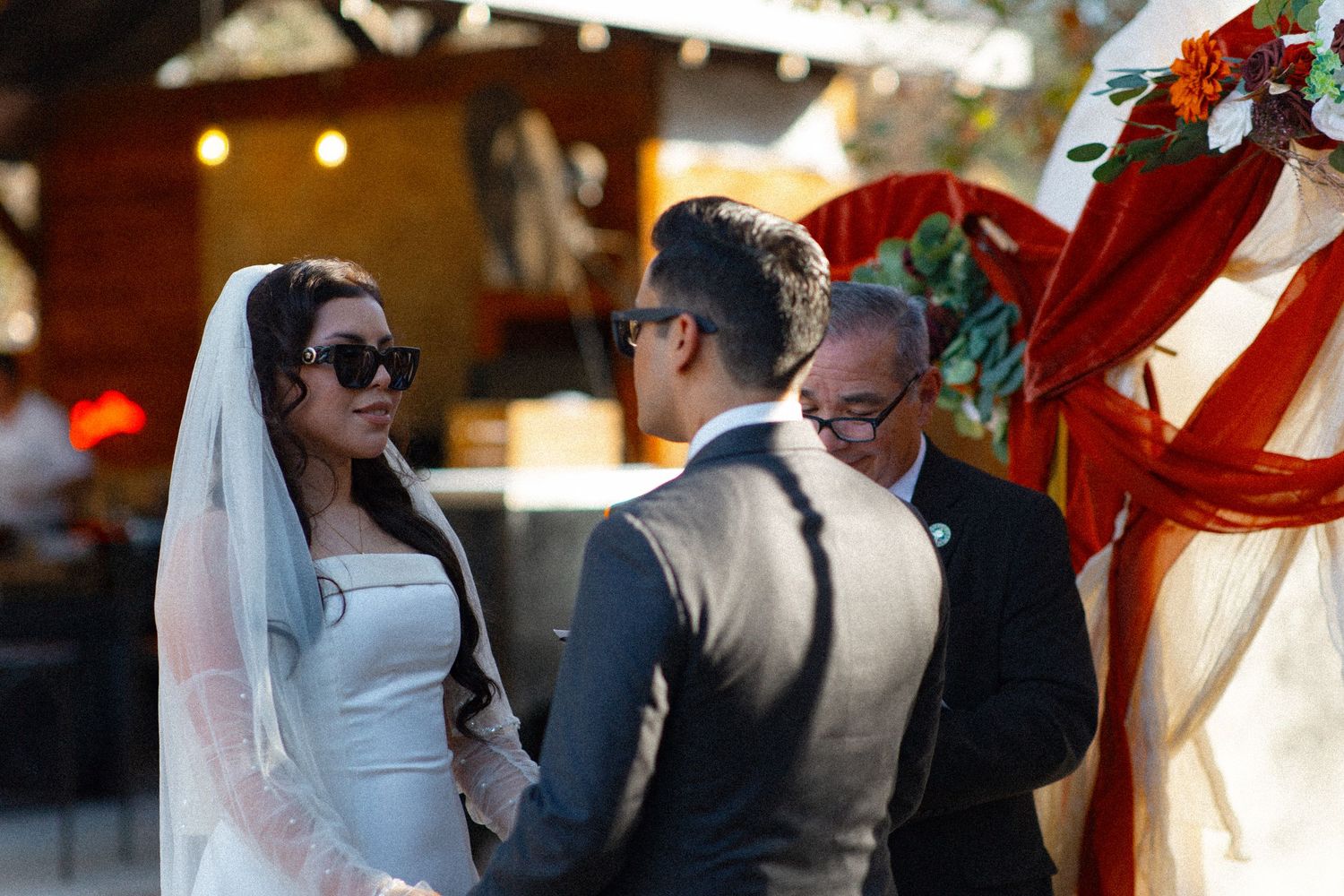 Outdoor wedding ceremony scene with couple exchanging vows under decorative red floral arrangements.