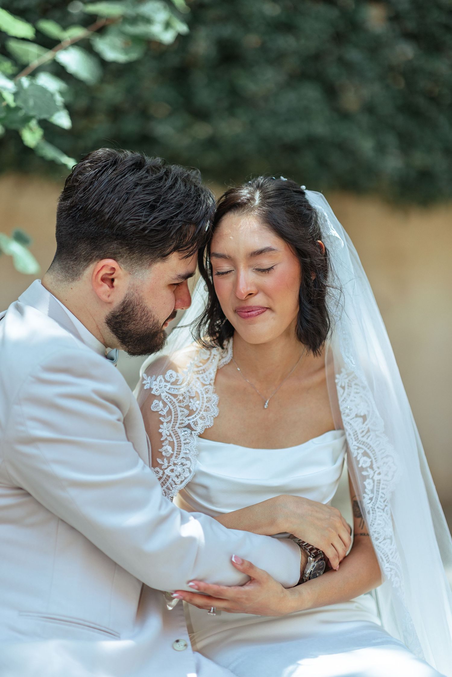 A romantic wedding portrait showing tender moments between newlyweds against green foliage.