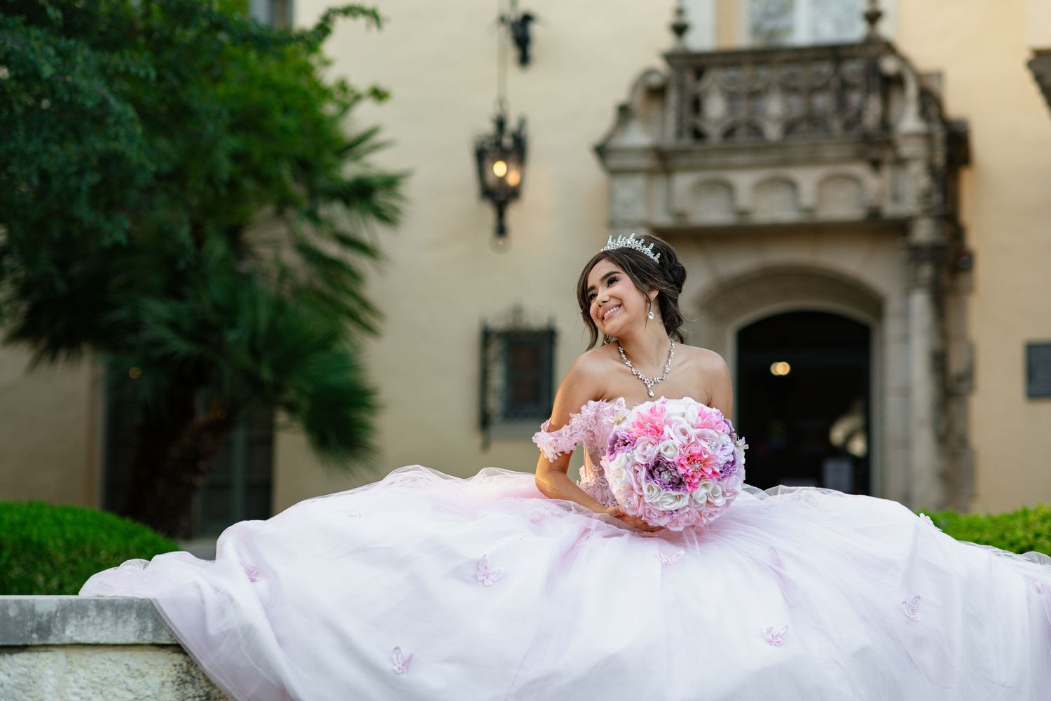 An elegant white ball gown with pink bouquet photographed on stone steps of a grand building.
