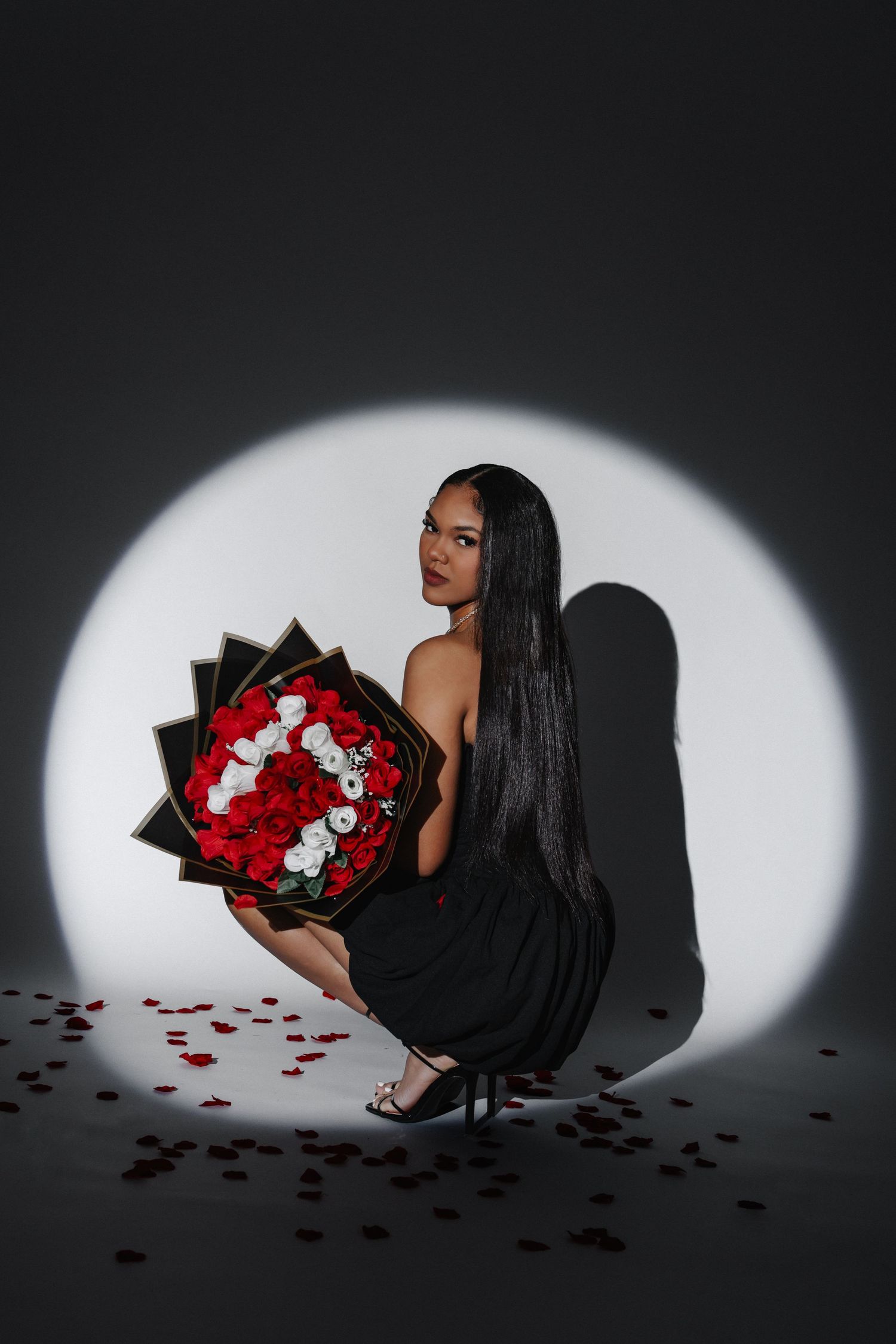 Artistic portrait of someone in a black dress holding a bouquet of red and white roses against a spotlight backdrop.