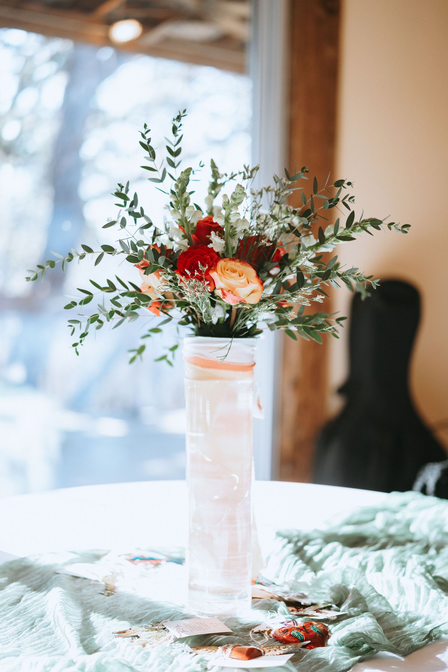 Delicate floral arrangement with red and peach blooms in frosted vase against bright window.