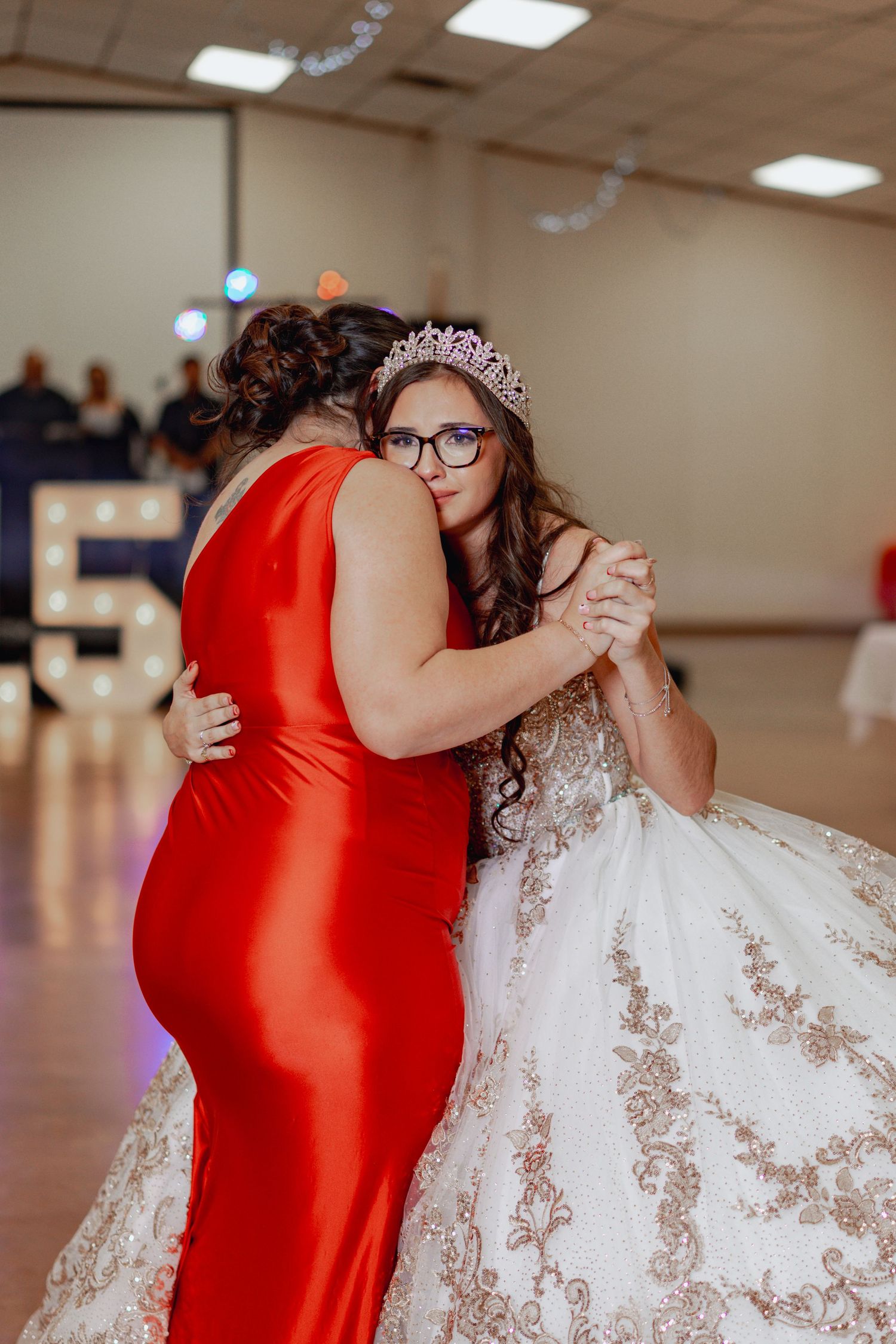 Two people share an emotional embrace at a celebration, one wearing a red dress and the other in an ornate white gown.