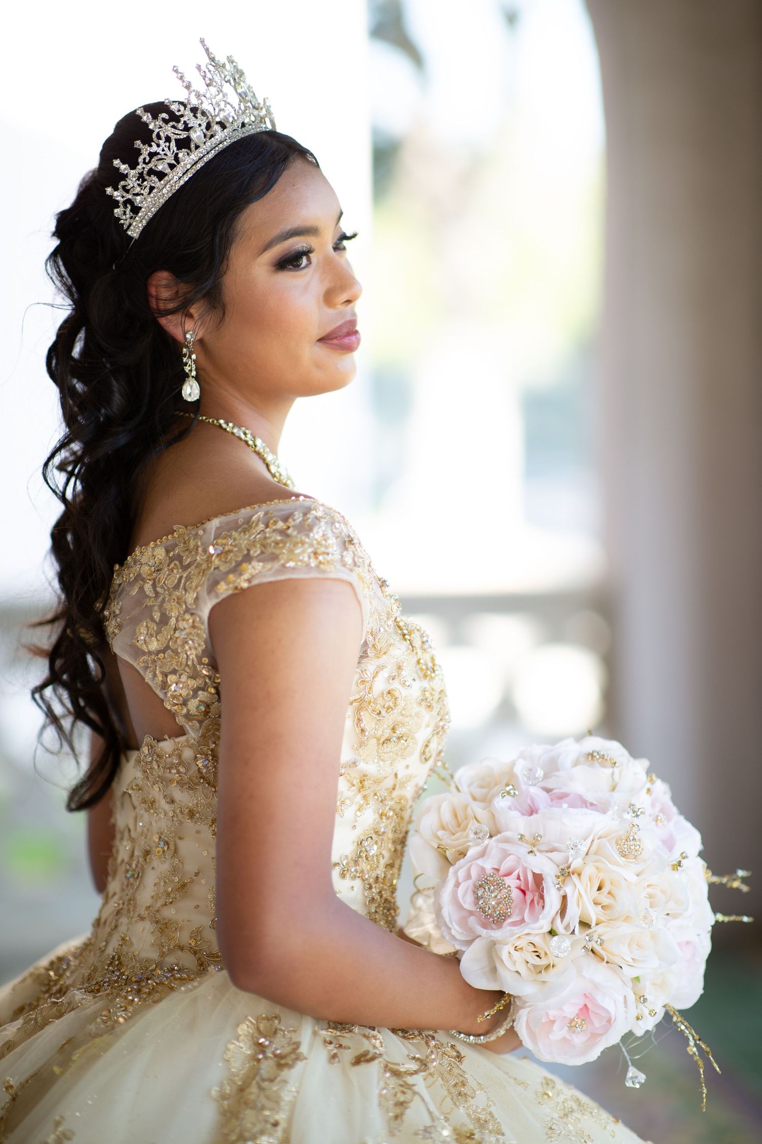 Elegant cream ballgown with gold embellishments and pink bouquet in natural light setting.
