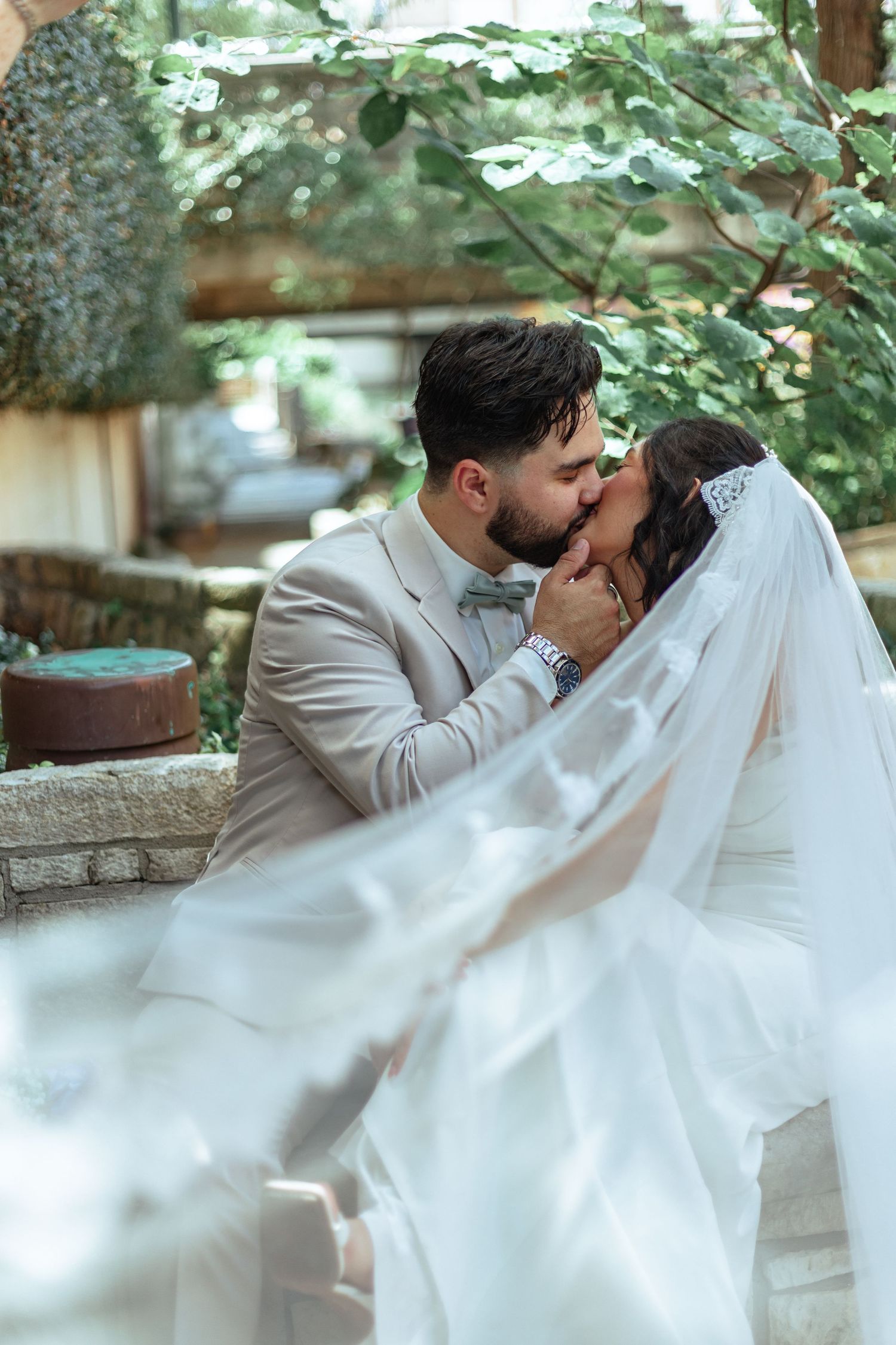 A romantic wedding couple sharing a tender kiss in an outdoor garden setting with flowing white veil and beige suit.
