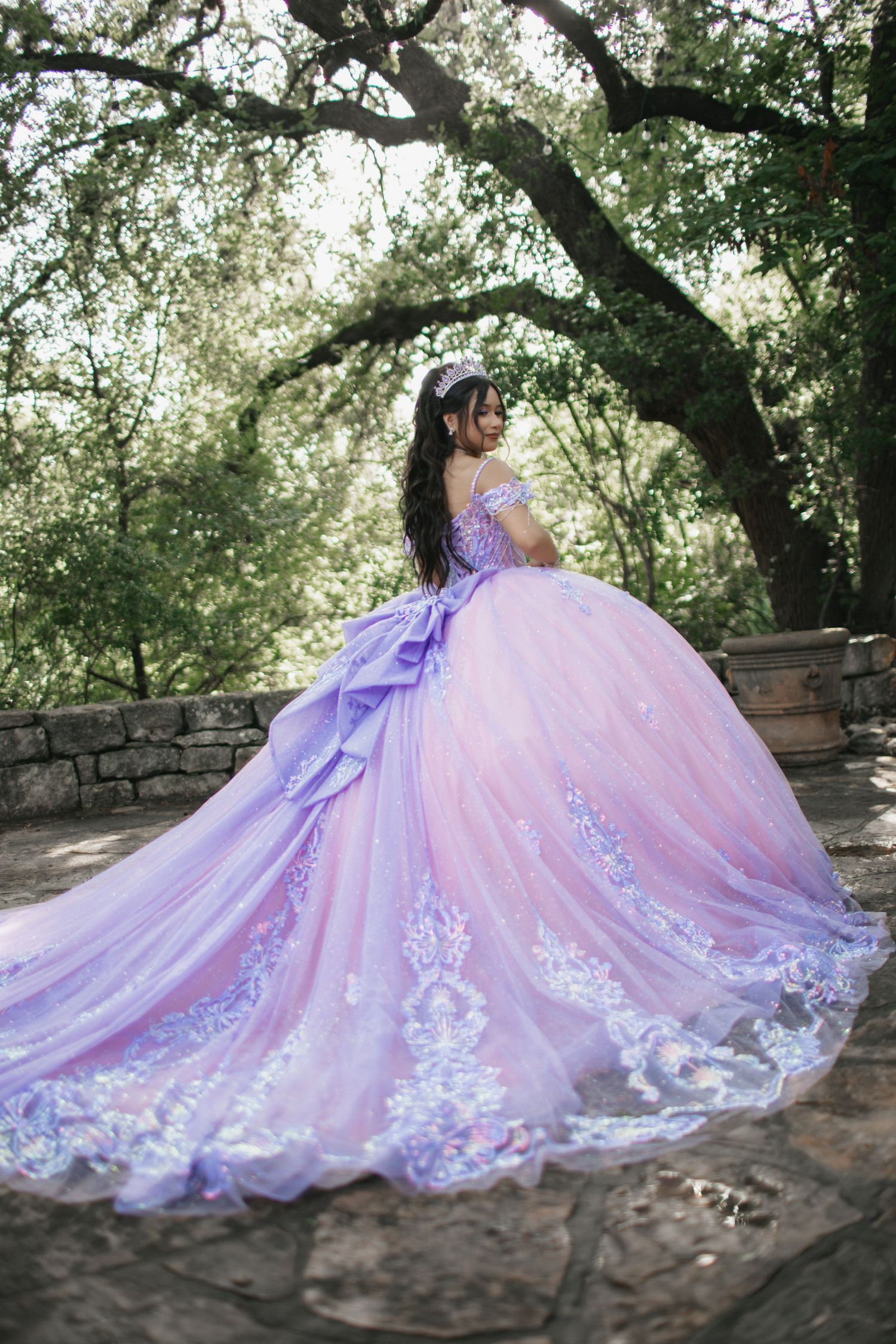 A lavender and pink ball gown with lace details photographed under a tree in a garden setting.